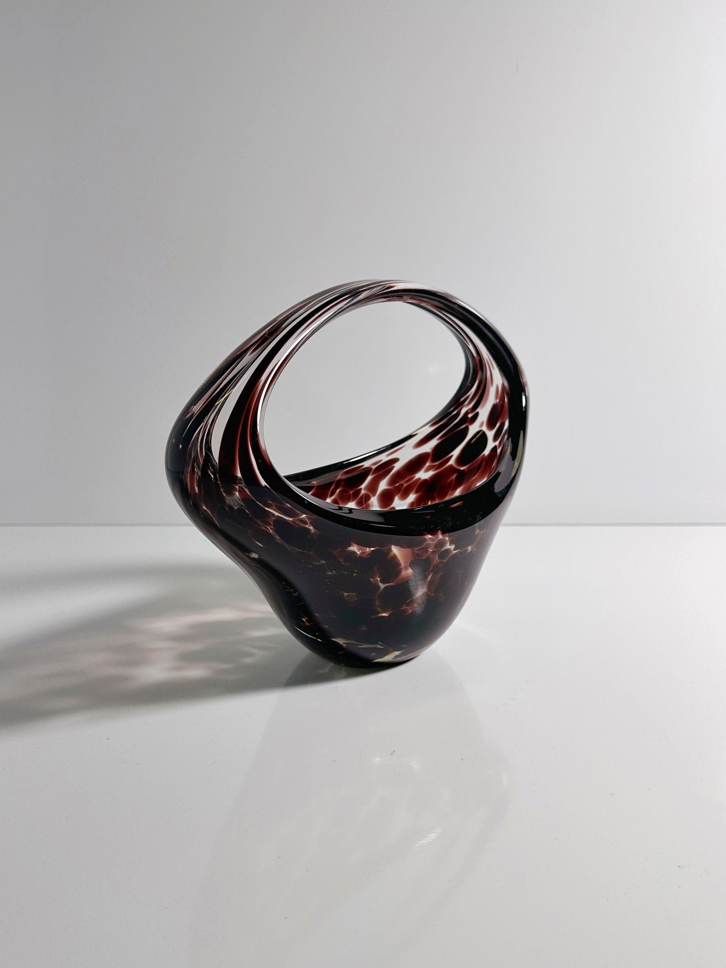 The Murano Tortoise Glass Handle Bowl from the 1970s by Dodo Vintage features an abstract, twisted design with deep red and brown translucent glass, showcasing swirling patterns. It stands elegantly on a reflective white surface against a plain backdrop.