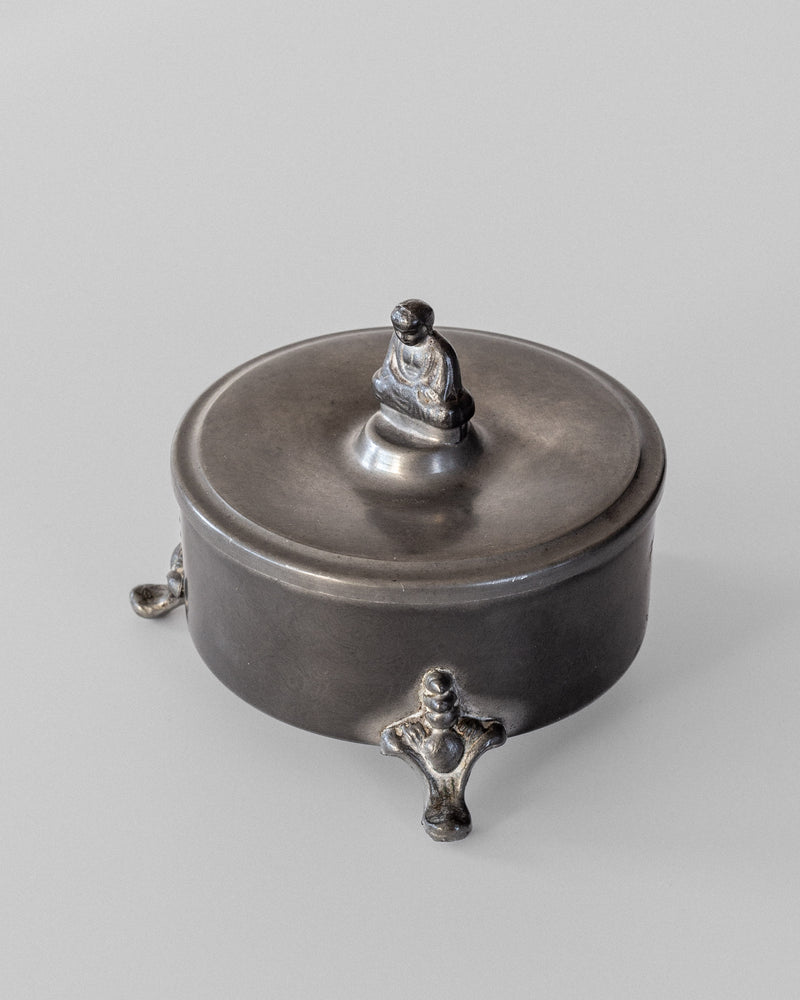 The Spigel Art Deco Metal Box from 1934 is a round, intricately designed container with a lid showcasing a small Buddha figure seated atop. Resting on three ornate feet, its sleek metal surface gently reflects light against a simple backdrop.