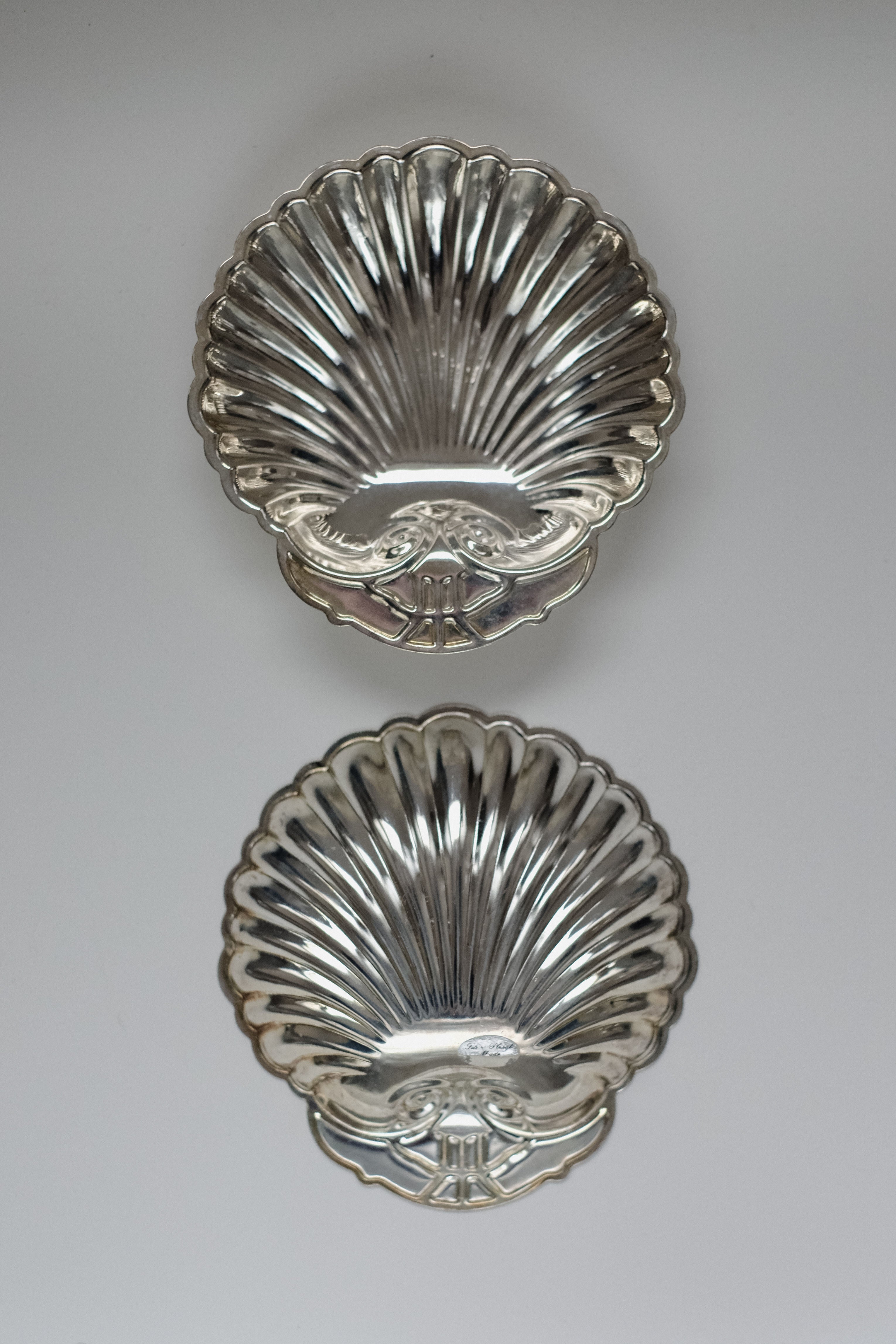 The Set of 2 Silver-plated Shell Trays by septembre studios features intricately detailed, ornate designs, adding timeless charm to any dining experience with their elegant, reflective surfaces against a plain background.