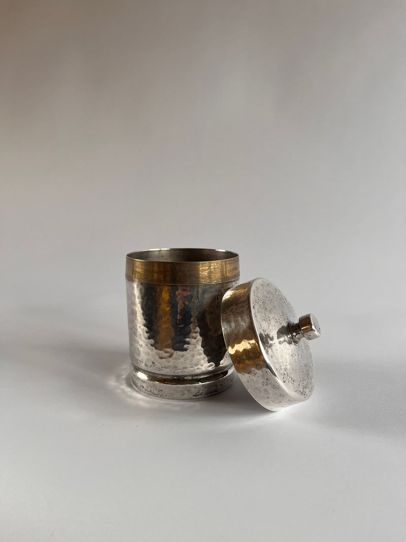 The Art Deco Hammered Pewter Box by Gero from Maud Vaughan is a textured, metallic cylindrical container with a matching lid. The shiny pewter surface on a neutral backdrop exudes elegant Art Deco charm.