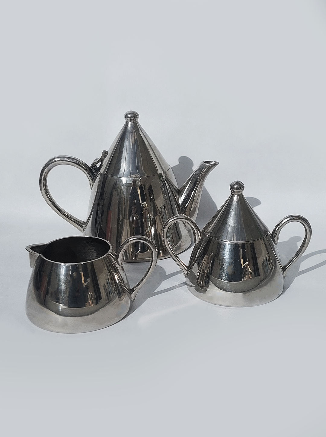 The Conic Modern Tea Set by Les Objoies features a shiny, brass silver tea set with a tall conic-shaped teapot and conical lid, accompanied by a small jug and a sugar bowl with a matching conical lid. Set against a plain, light background, this collection reflects light beautifully, highlighting its modern and minimalist design.