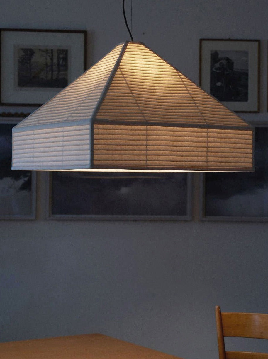 A Nassi Lamps Pyramid Hanging Lamp emits a warm glow illuminating a wooden table below. Behind, blurred black and white photographs are framed on a dark wall. A wooden chair is partially visible to the right of the table with a metal spring on top.