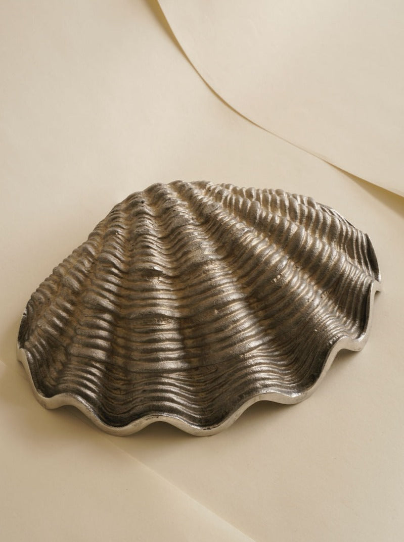 A silver, clam-shaped decorative serving dish featuring intricate ridges and grooves rests on a beige surface. The detailed texture mimics the natural contours of a clam shell, adding a sophisticated and elegant touch to the design. This is the Shell-Shaped Fruit Bowl by Les Objets Amsterdam.