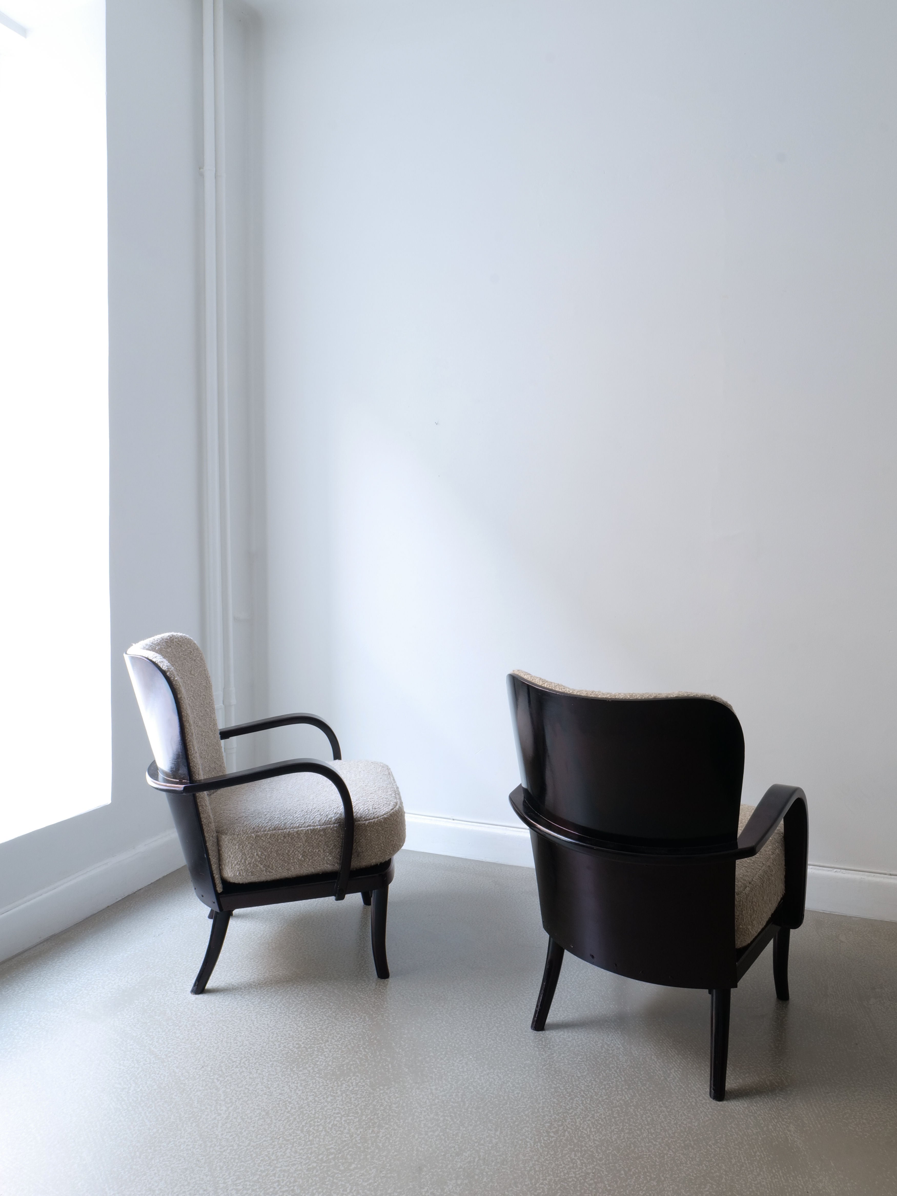 A pair of Werner West Armchairs from the 1930s, crafted by Collection apart with sleek black lacquered wood frames and upholstered in Moretti bouclé, are positioned near a spacious, bright window in a minimalist room featuring white walls and a light-colored floor. The armchairs face each other, fostering a cozy yet open ambiance.