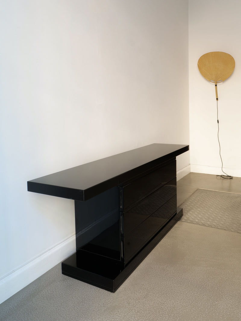 A sleek and modern High Gloss Console Table 1970s from Collection apart, finished in black, stands elegantly against a white wall on a polished concrete floor. A minimalist beige wall lamp with a fan-like design is partially visible on the right side, enhancing the custom-built elegance of the scene.