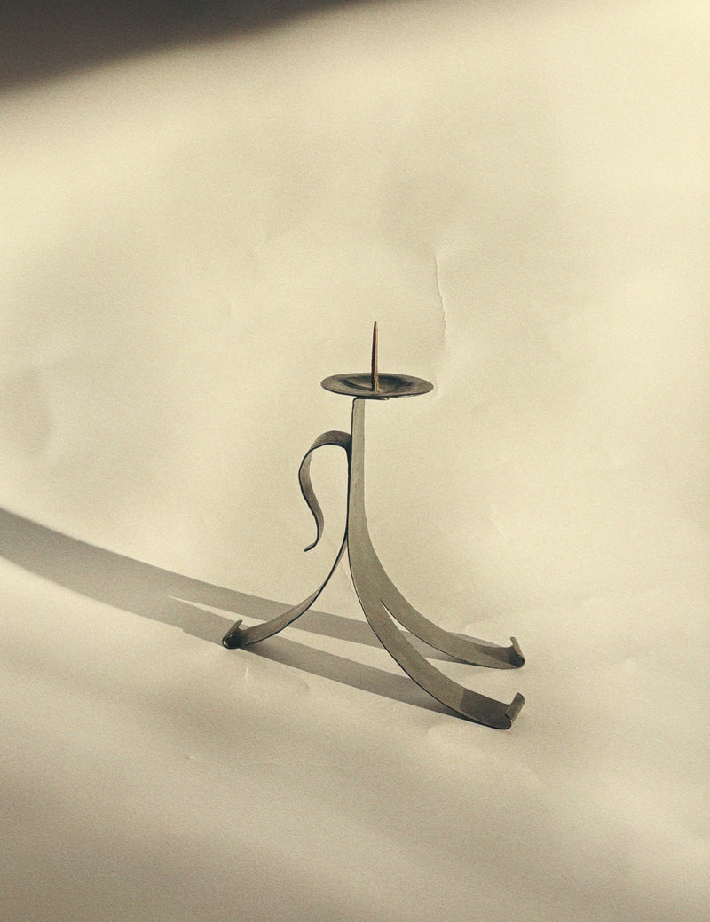 A handmade metal candleholder by Huerto, featuring a sleek, curved design, sits on a sunlit surface. This skillfully crafted piece of artist-made tableware casts a shadow on the light background, highlighting its artistic simplicity and vintage charm.