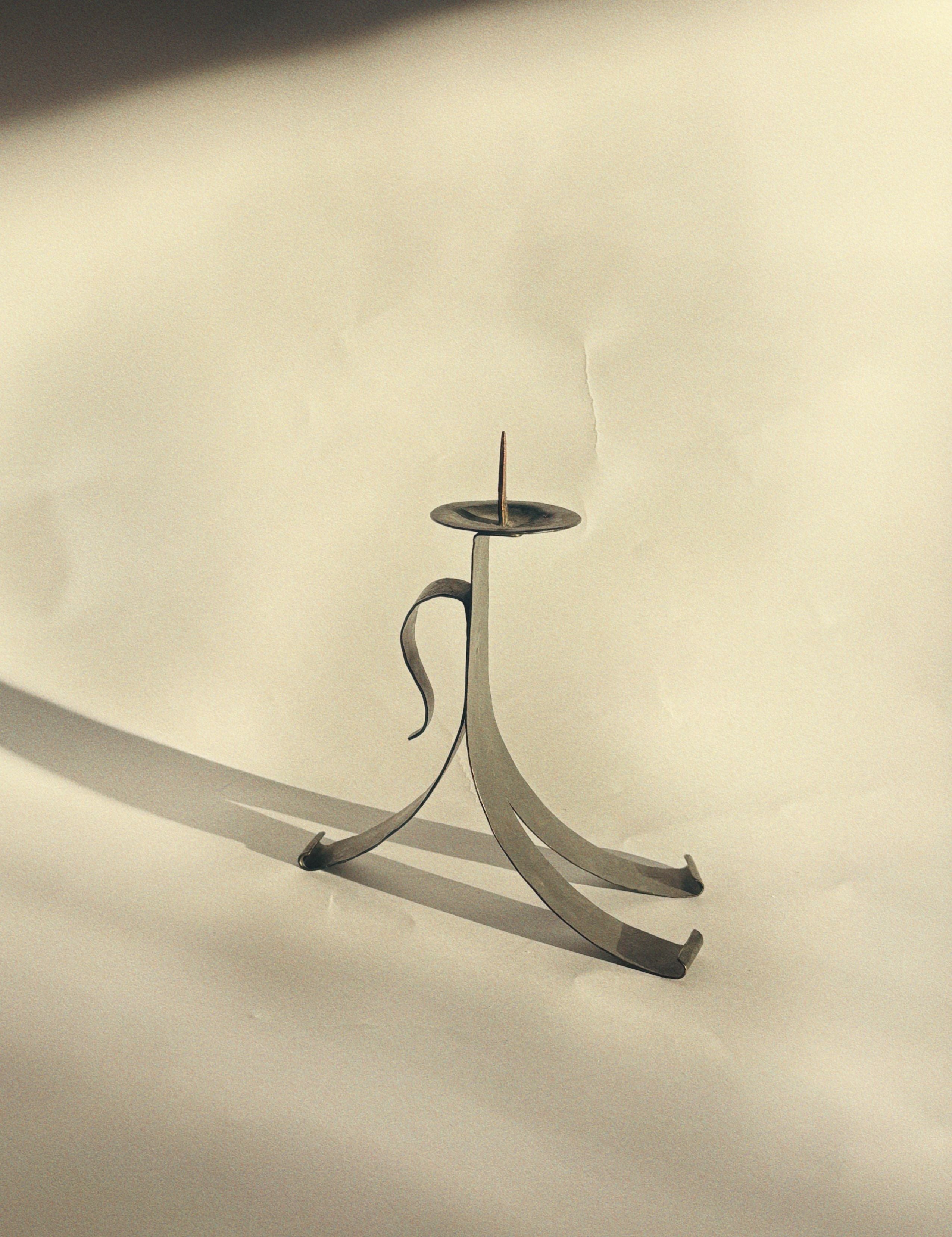 A handmade metal candleholder by Huerto, featuring a sleek, curved design, sits on a sunlit surface. This skillfully crafted piece of artist-made tableware casts a shadow on the light background, highlighting its artistic simplicity and vintage charm.