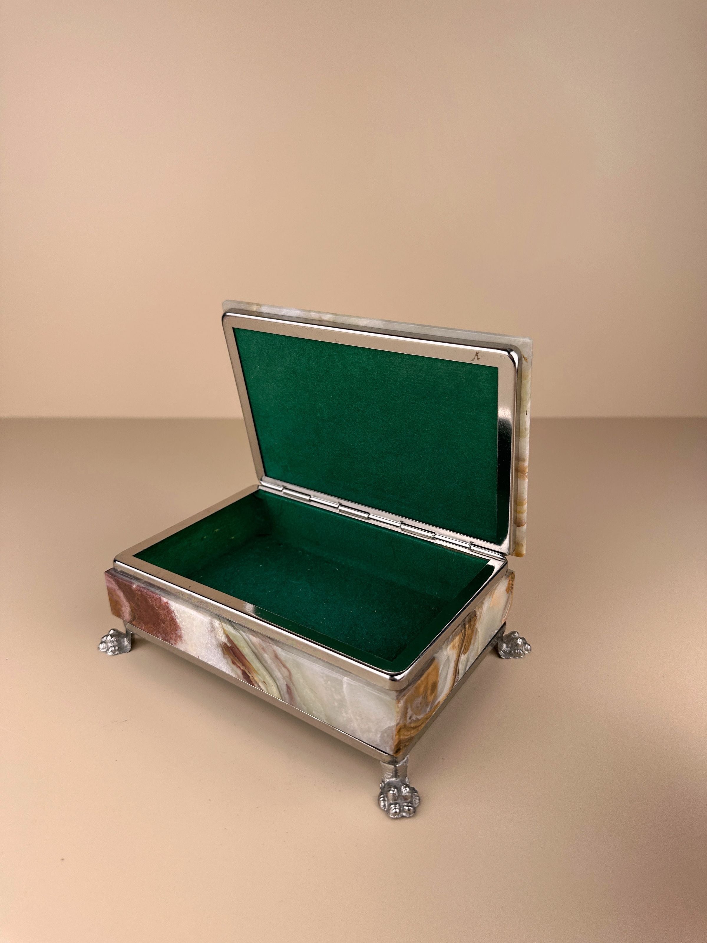 A close-up photo of a 1970s Italian onyx jewellery box by Dodo Vintage with a silver exterior, green felt interior, and four ornate feet. The elegant box is positioned against a plain beige background, showcasing its timeless design.