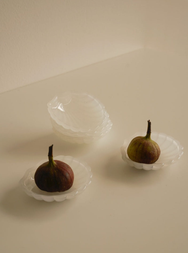Four small, white, shell-shaped Les Objets Amsterdam 5x Shell Dessert Plates are arranged on a light surface. Two plates each contain a ripe fig, while the other two delicately crafted plates are stacked and empty. The scene exudes a minimalist and elegant aesthetic.