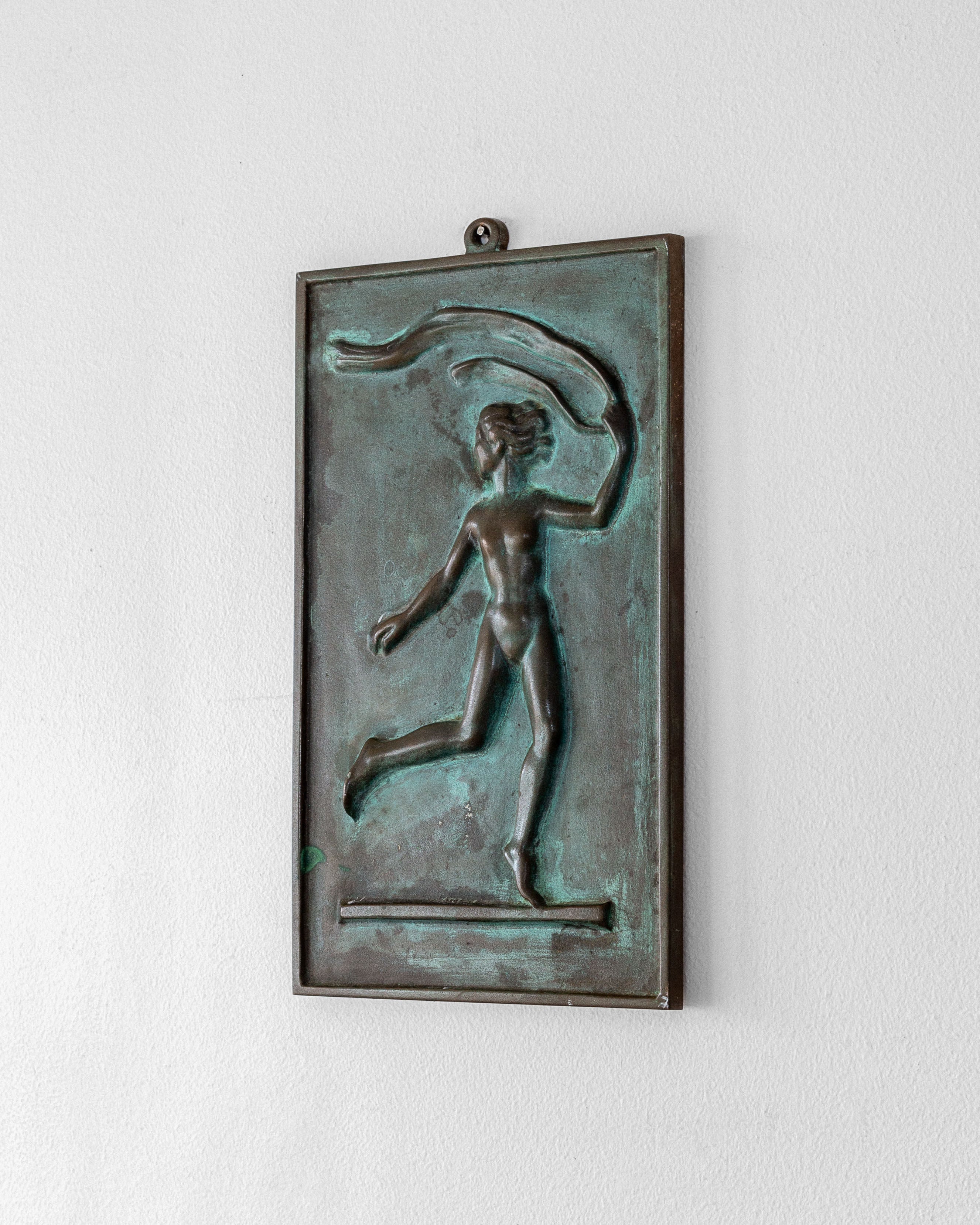 The Bronze Wall Relief by David Jahrl from Spigel features a dancing figure with a flowing ribbon on a rectangular plaque, epitomizing Swedish Grace and elegantly displayed against a plain white wall.