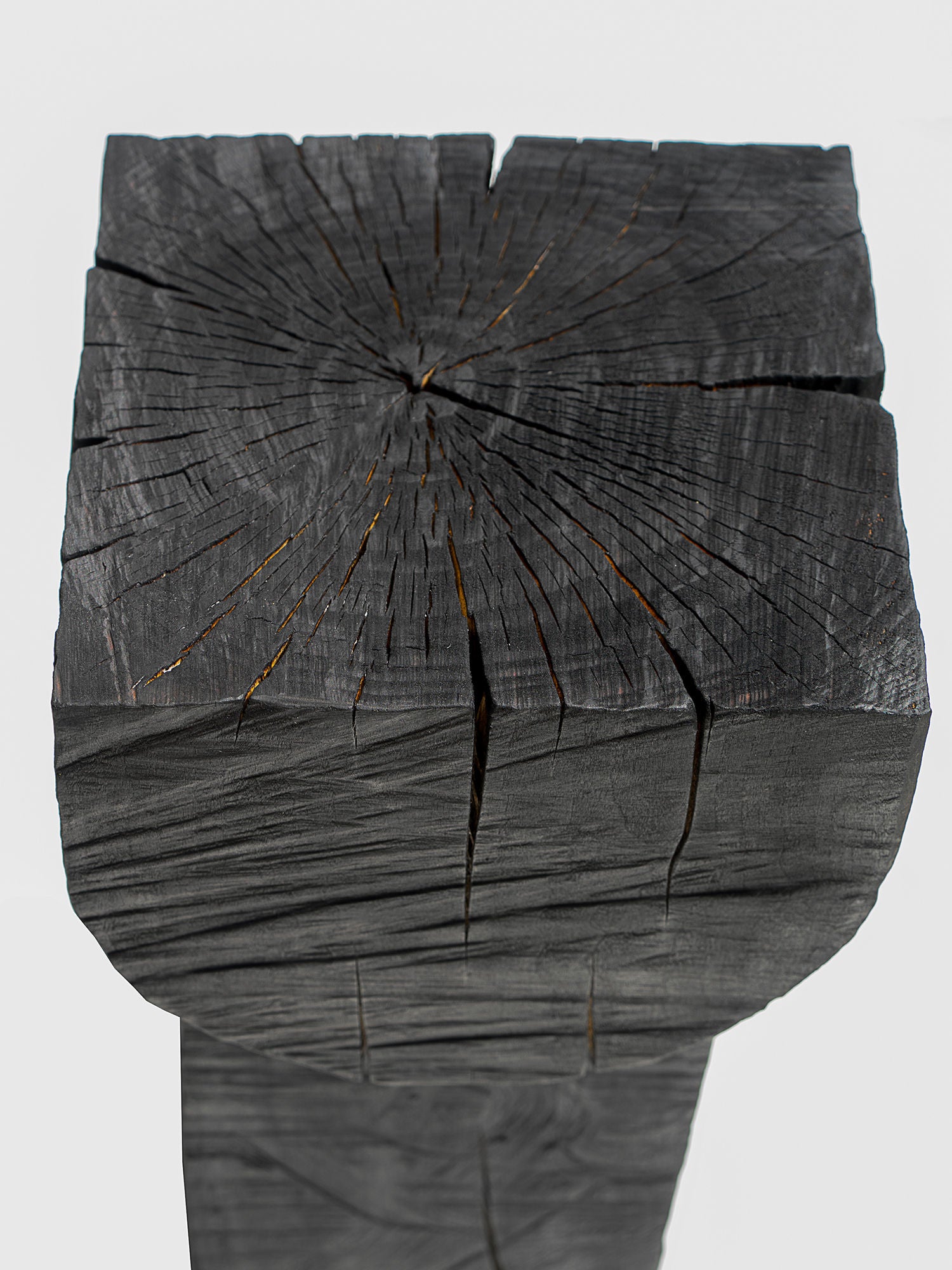 The Burnt Black Coffee Tables/Stools by Touch With Eyes feature a charred wooden design with natural cracks and a rough texture. Radial splits on the top surface and visible wood grain along the side provide a striking contrast against a light gray background.