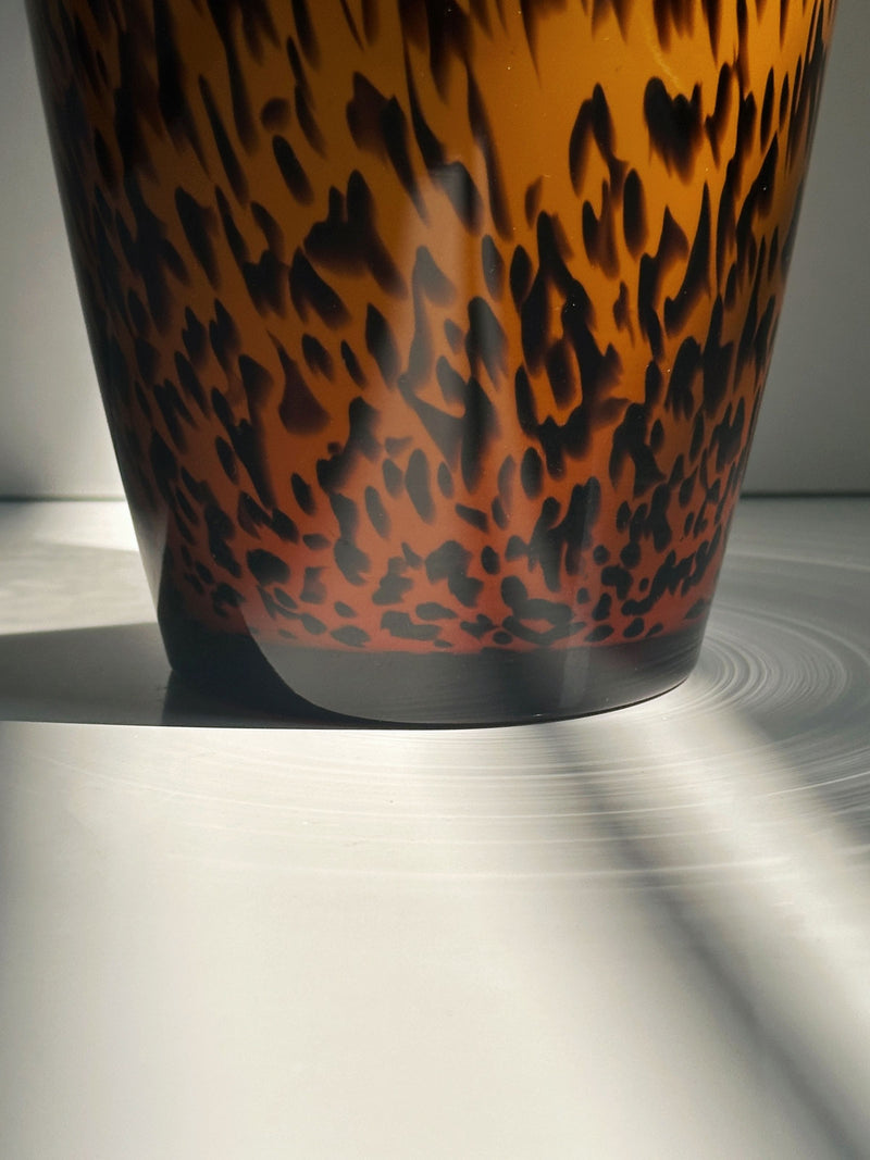 A close-up of the 1980s Murano Vase Tortoise pattern by Dodo Vintage, featuring a round shape with brown and black design, sits on a white surface with sunlight casting shadows and reflections.