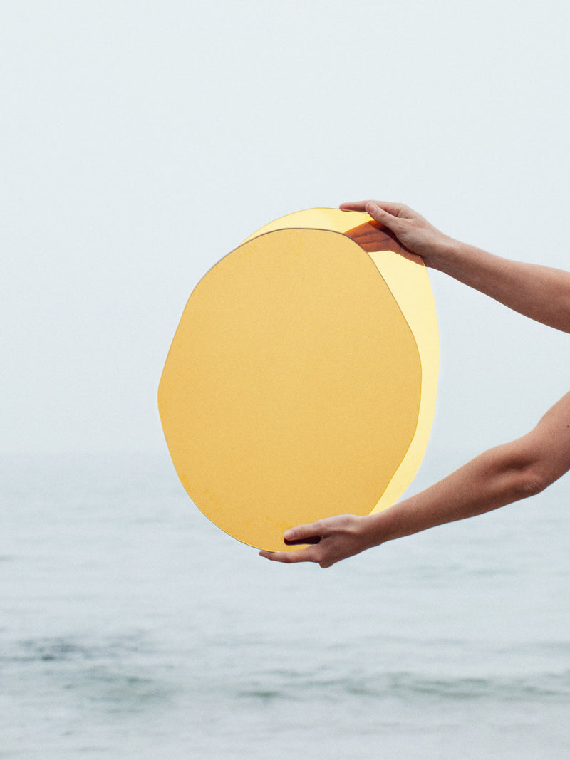 A person is holding a Mirror Ombrée Gold by Laurène Guarneri with both hands extended in front of a hazy seascape backdrop. The light gray sky and calm water create a serene atmosphere, reflecting off the coloured glass like a poetic mirror of nature’s tranquility.