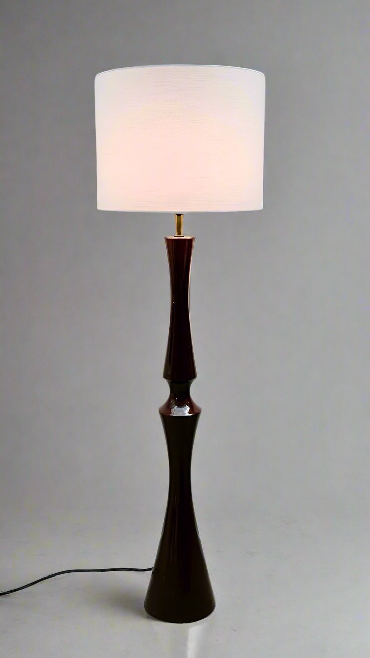 A tall Lampadaire en céramique attribué à Cari Zalloni with a dark brown, intricately designed base and a white cylindrical lampshade stands against a plain white wall, emitting a warm, diffuse light. Designed by LA NOUVELLE GALERIE, this elegant piece features a black power cord visible at its base, lying on the light-colored floor.