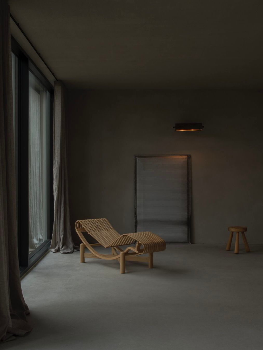 A minimalist room showcases a Bicci de' Medici wooden lounge chair inspired by Jean Prouvé, paired with a small stool against dark gray walls. Soft light filters through floor-length curtains, complementing the subtle wall-mounted light fixture for understated illumination.