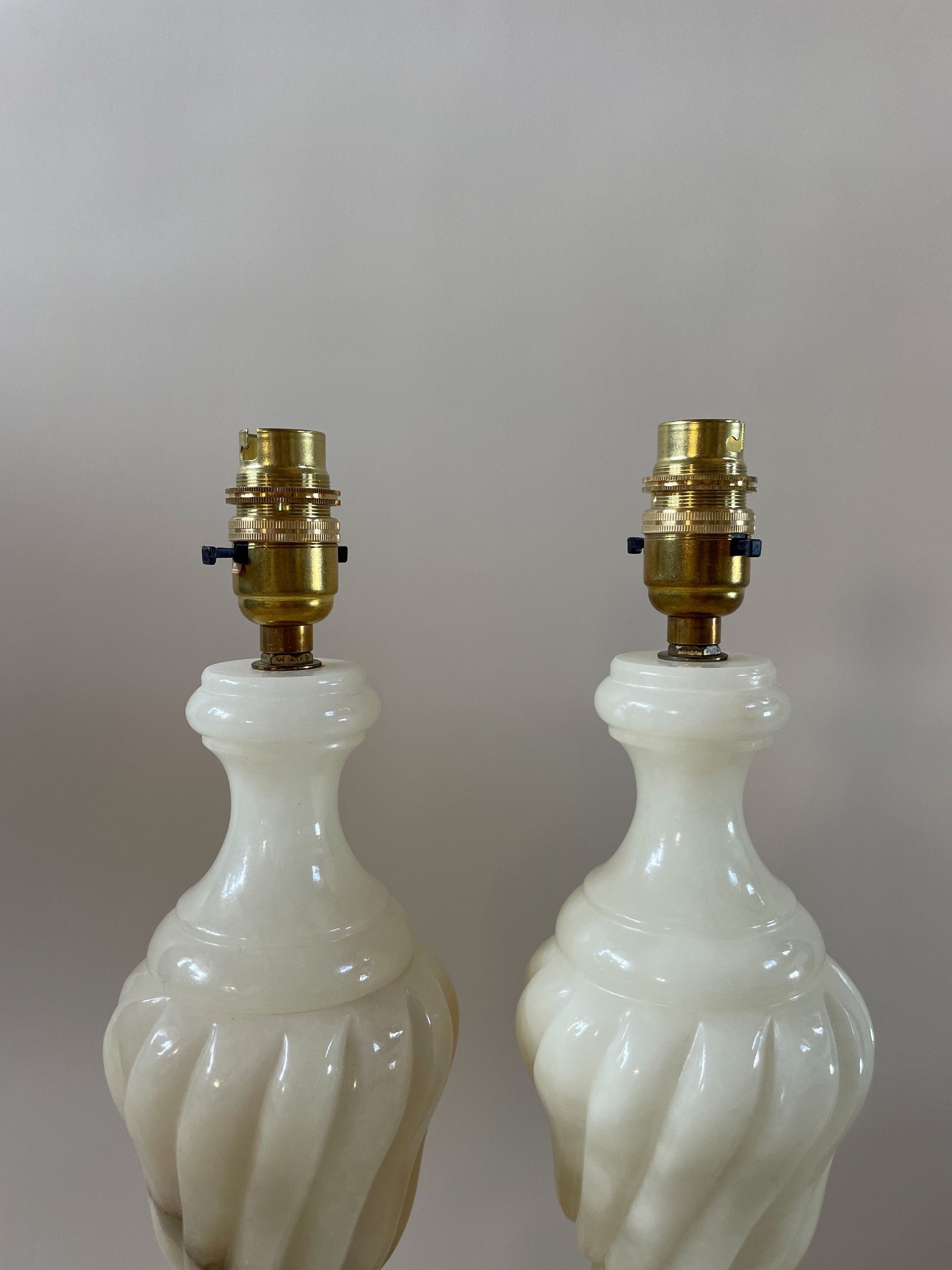 Two vintage alabaster lamps from Maud Vaughan feature off-white, swirled ceramic bases. These decorative lamps have been rewired with antique gold flex and come without shades, highlighting their brass bulb holders and electrical fittings at the top. They are displayed against a plain, neutral background.