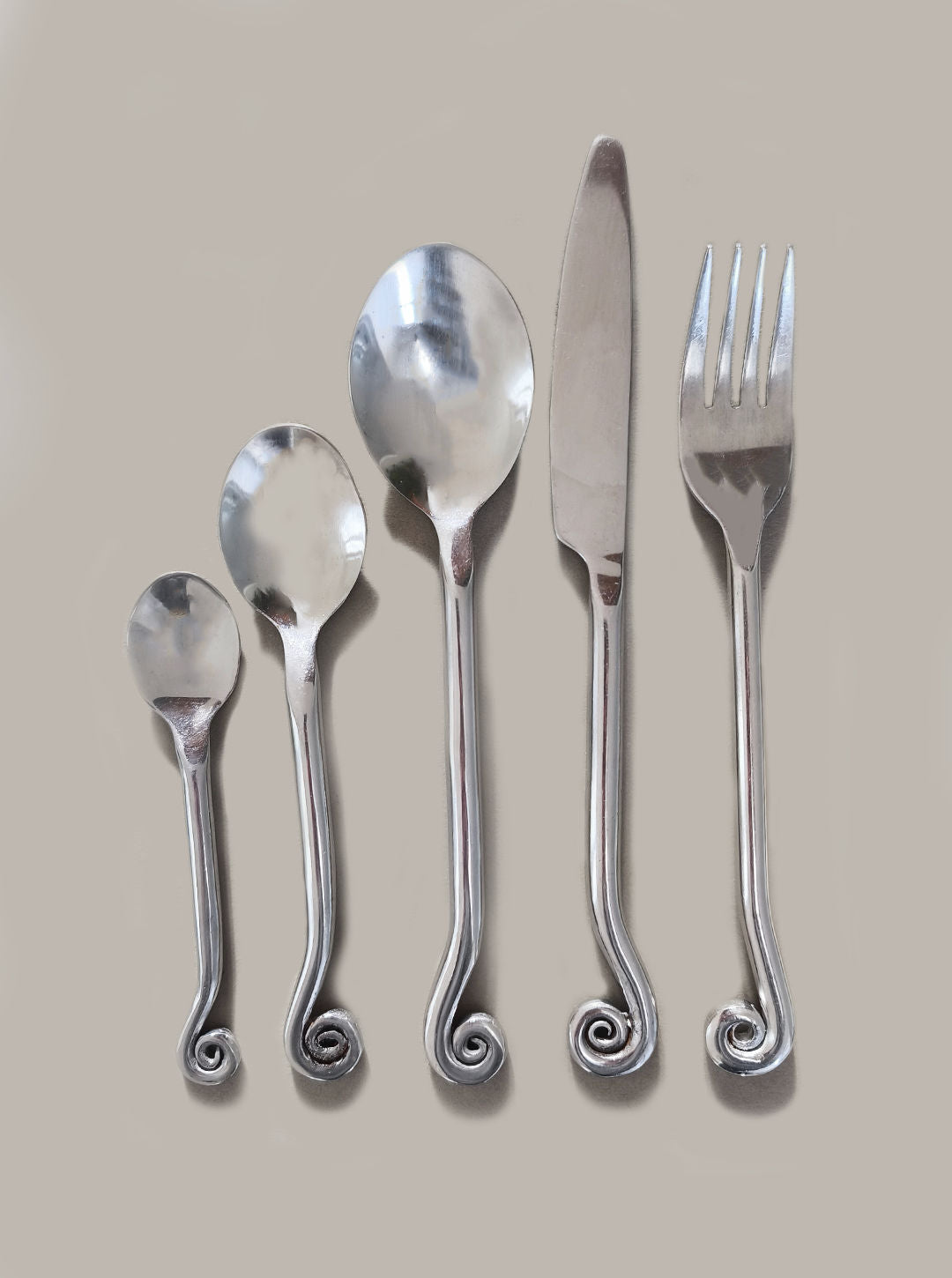 Les Objoies presents the Set of 5 Swirling Cutlery: a contemporary silver set including a small spoon, larger spoon, soup spoon, knife, and fork, all stylized with unique spiral-ended handles on a gray background.