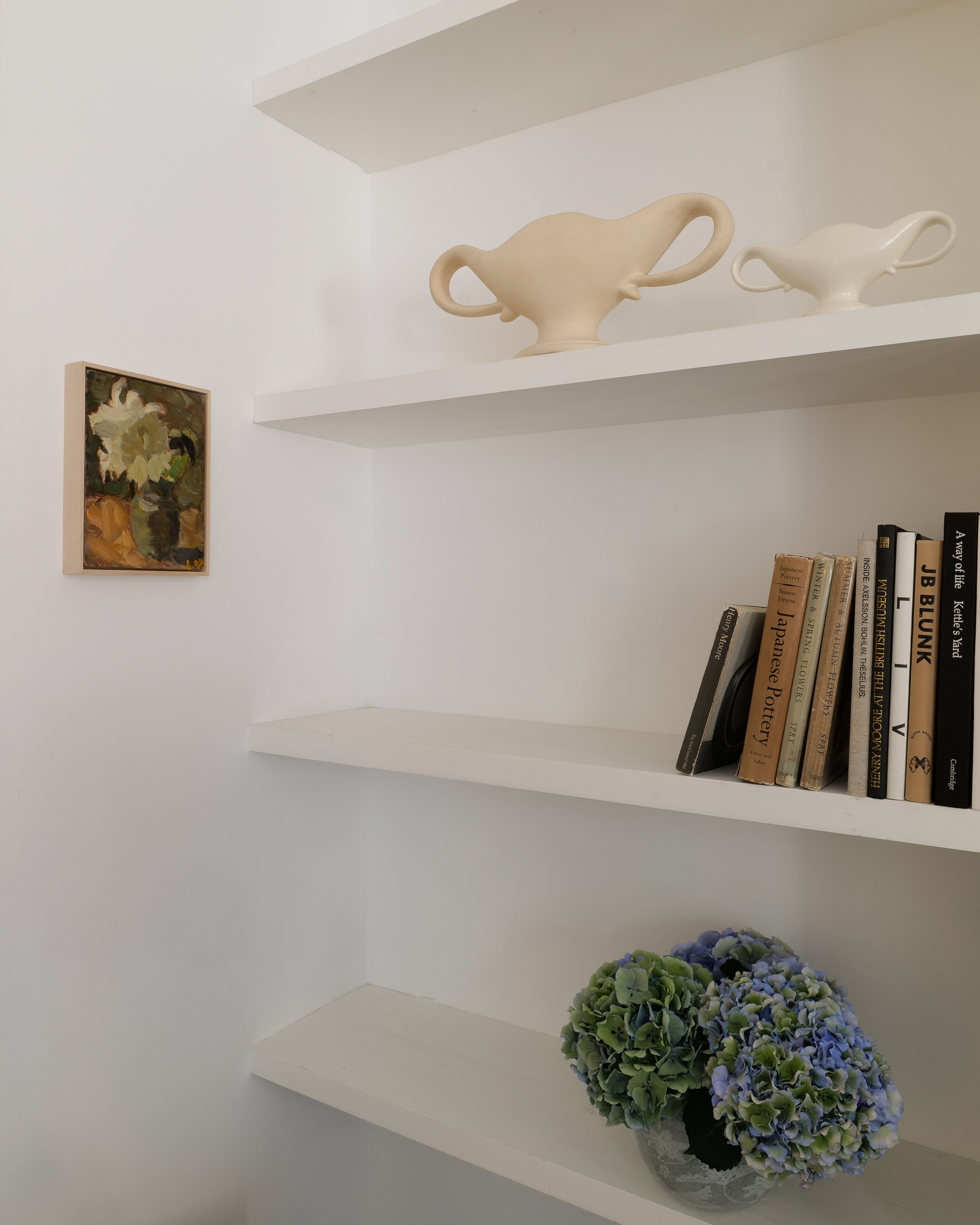 A minimalist white shelving unit by Lars Vidlund from Collection apart showcases a small still life painting, two vases with organic shapes in beige and white, a collection of books, and a bowl of blue hydrangeas. The shelves and overall setting emphasize simplicity and aesthetic decor in an autumn color palette.