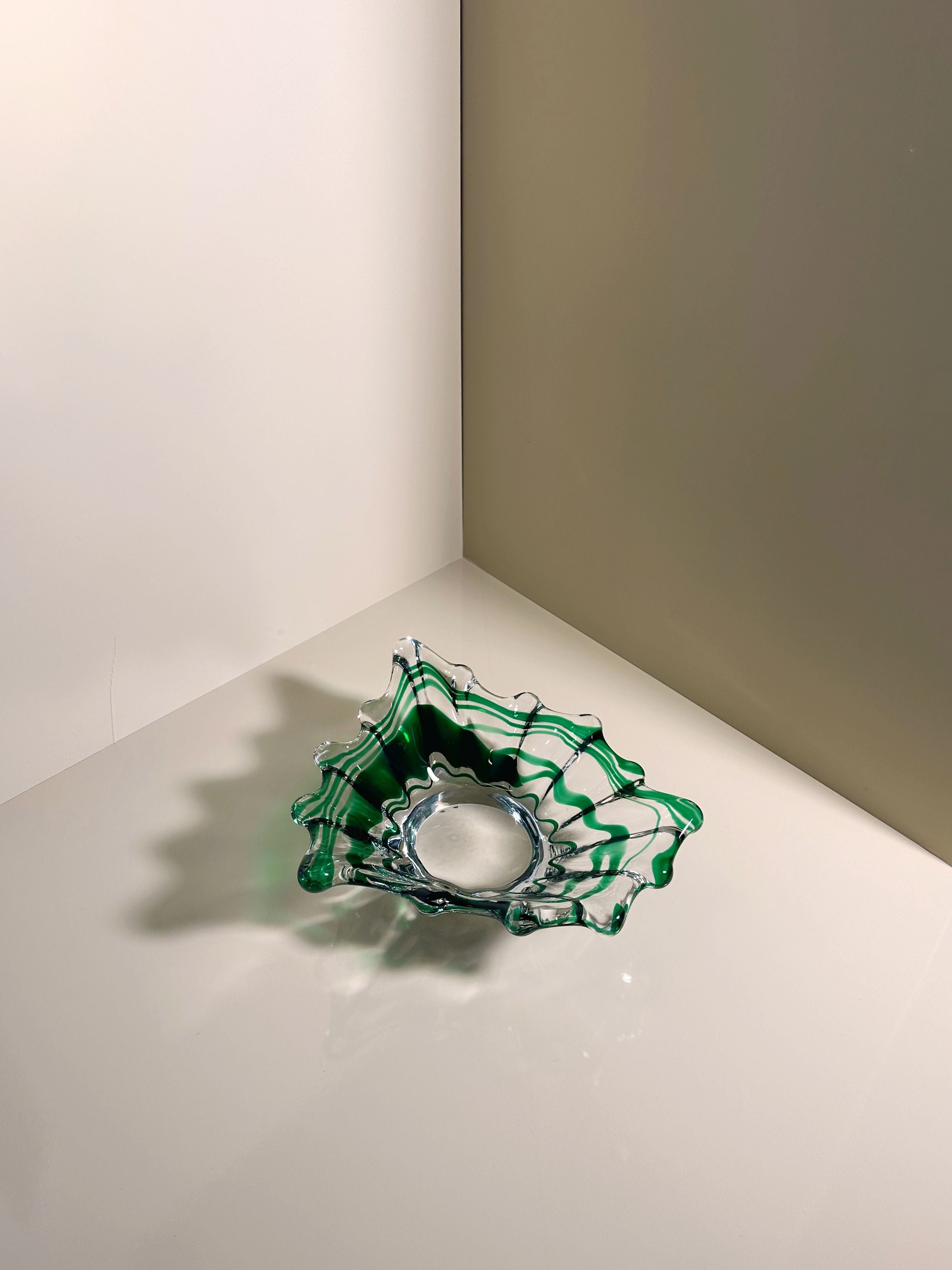 In the room's corner, a green swirled Murano vide-poche by Dodo Vintage rests on a glossy surface. Soft lighting casts delicate shadows, accentuating its elegant curves and intricate design.