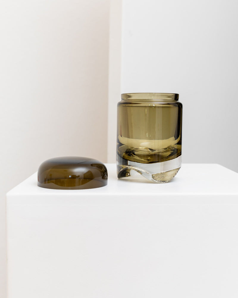 A small Vintage Glass Jar 1970s by Bottega Jacobs, featuring a translucent, olive-green hue, is displayed with its cylindrical lid beside it on a white pedestal against a neutral background. The jar exudes mid-century design elements with its smooth cylindrical form and slightly domed lid.