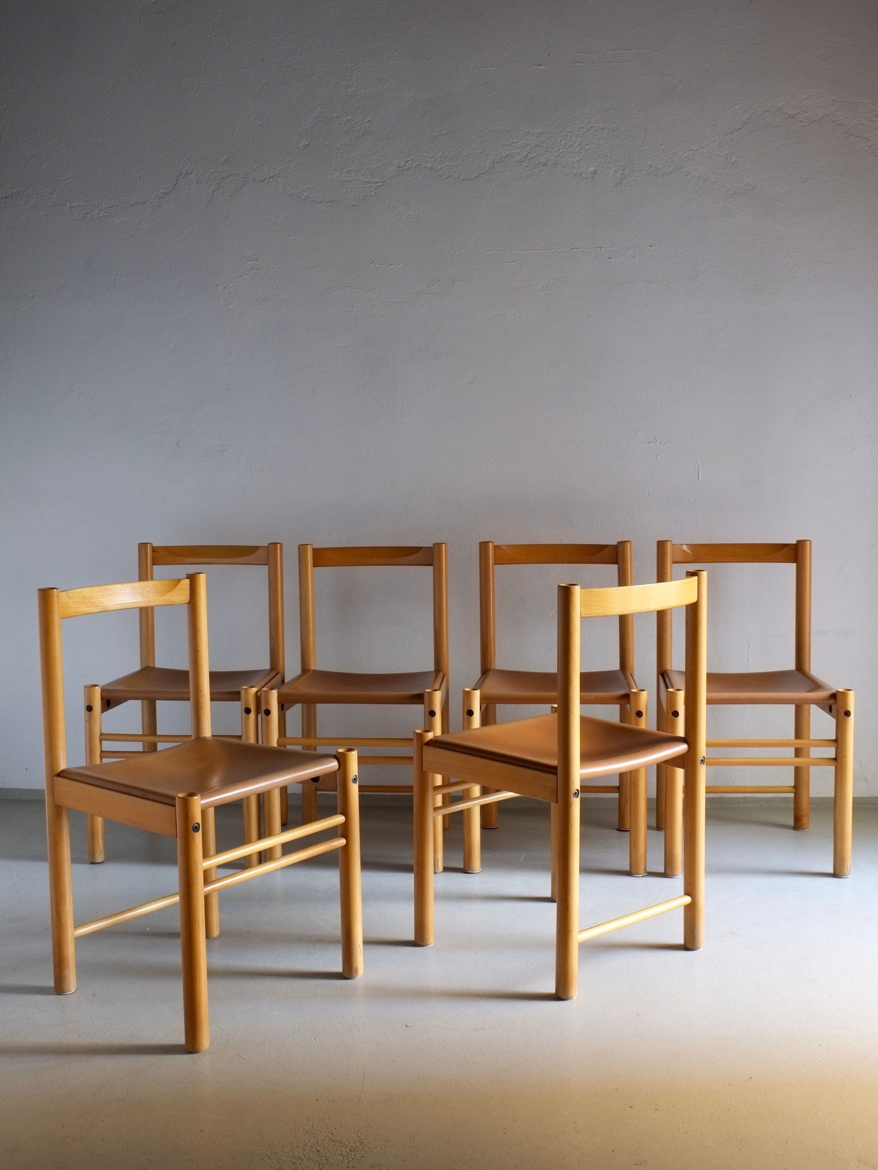 Six Beech Wood Cognac Leather Chairs from Ibisco, Italy, 1960s by Veter Vintage are arranged in two rows against a light gray wall on a smooth floor. The light highlights their beech wood finish and cognac leather seats, creating a warm and inviting atmosphere with hints of vintage charm.