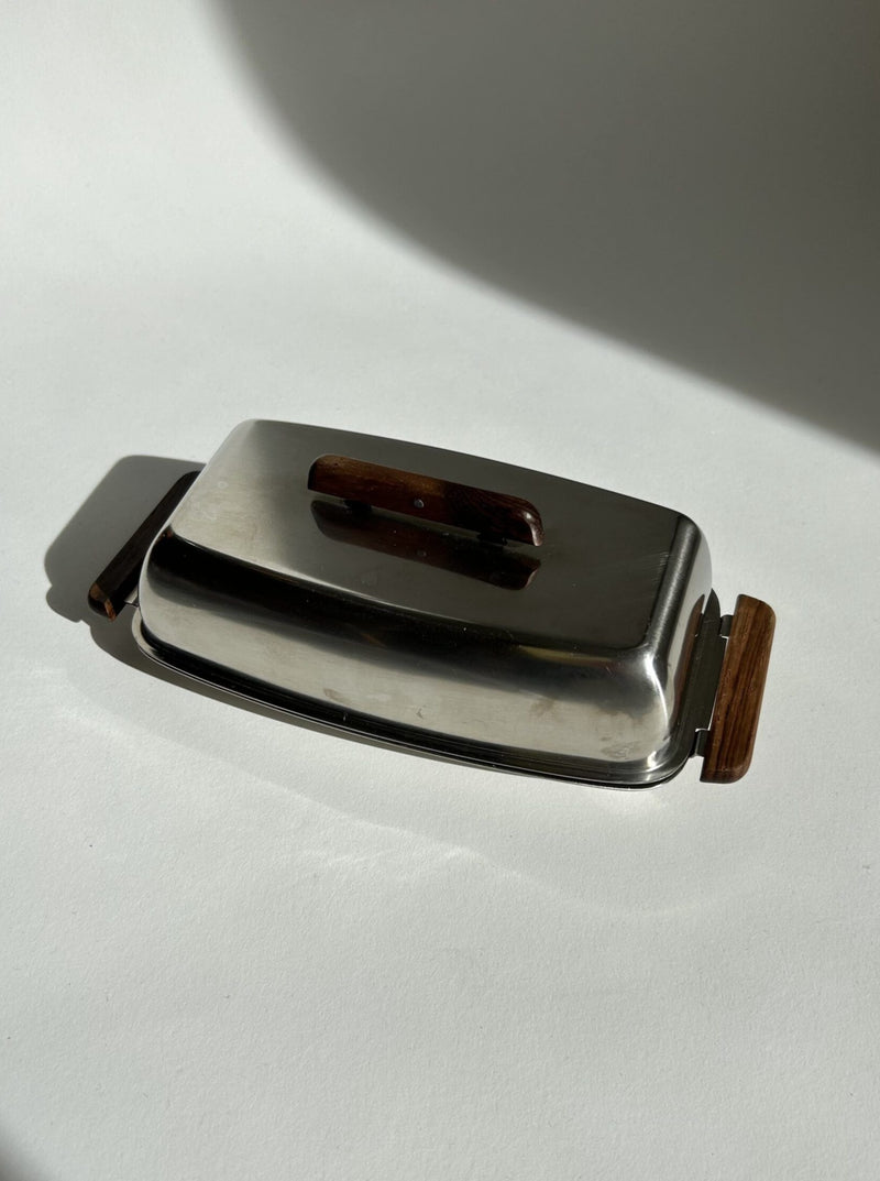 The Mid Century Butter Dish by Stelton, available at Flock Studio, exudes vintage charm with its polished stainless steel and wood finish. Its mid-century design, featuring wooden handles and sides, casts an elegant shadow on the light surface, highlighting its timeless appeal.