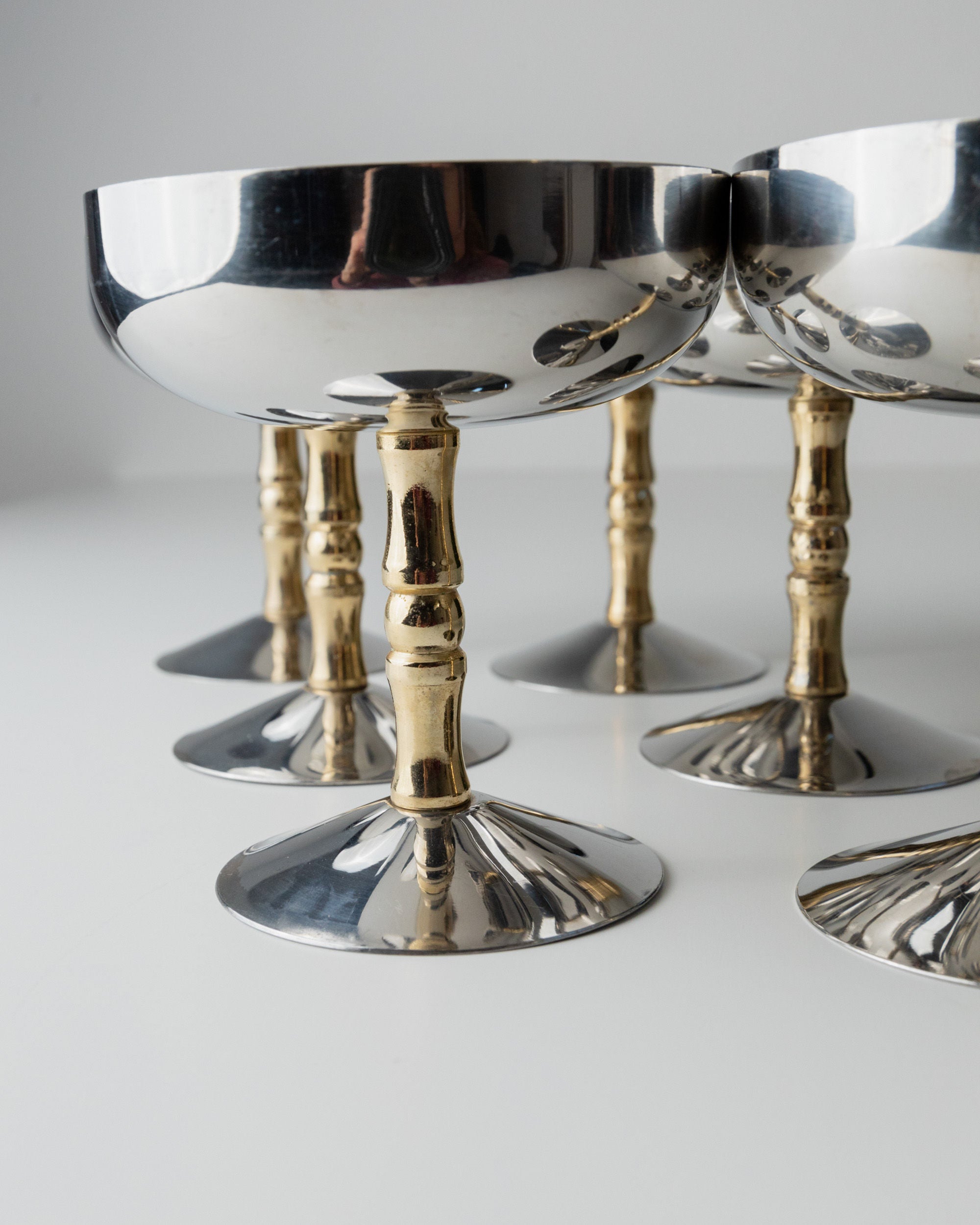 On a white surface, six Bottega Jacobs 6 Stainless Steel Champagne Coupes 70s showcase reflective metal bowls and gold-colored stems, blending sleek modern design with vintage stainless steel aesthetics for classic sophistication.