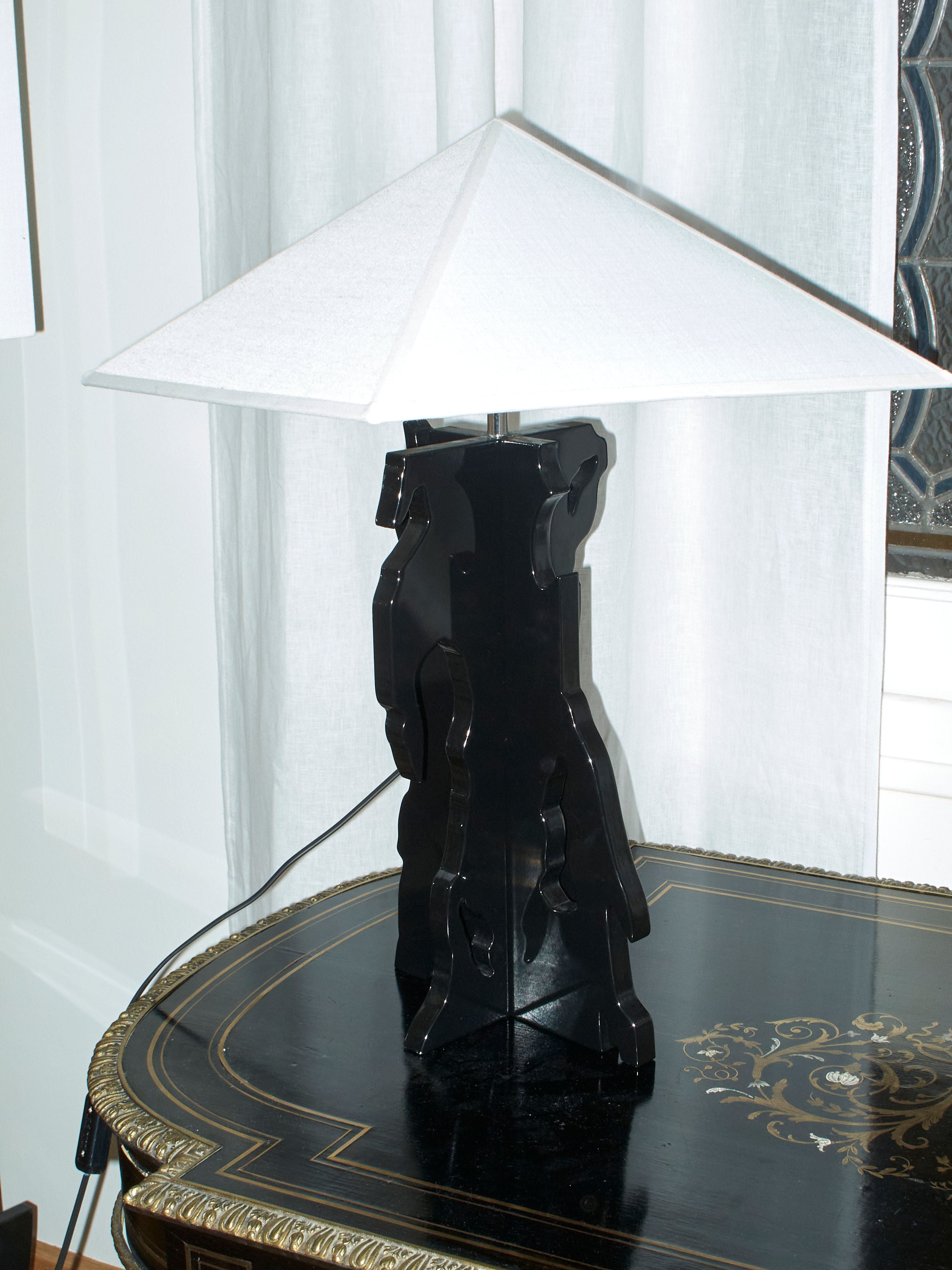 The Bird" Table Lamp by Ochoa Rocabert, featuring a white, angular shade and an abstract base, sits on a decorative black table with gold accents. Its design captures the modern aesthetic of its surroundings as an engaging conceptual piece. White curtains hang in the background, enhancing the chic ambiance.