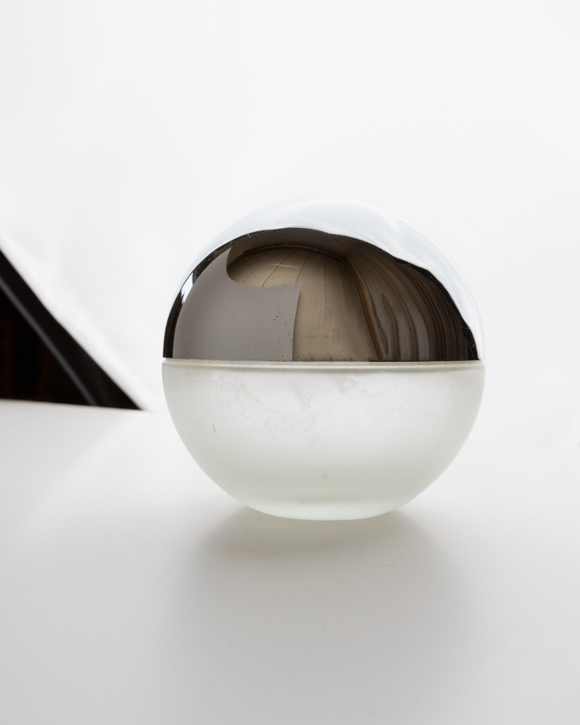 A Liqueur Bottle from Bottega Jacobs, Italy 1980s, with a chrome-like top and clear glass bottom sits on a white surface. It reflects abstract shapes and light against a bright background, evoking a space-age design for a minimalistic, modern composition.