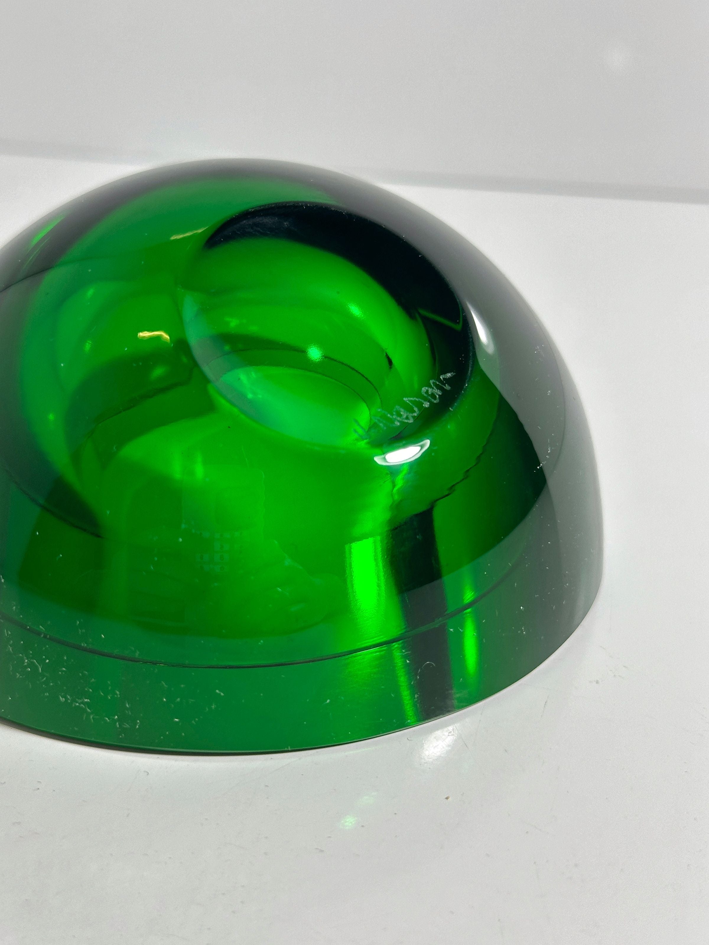 The Dodo Vintage 1960s Space Age Glass Ashtray features a smooth, translucent green dome on a white surface, reflecting light and casting shadows. Its glossy finish showcases craftsmanship with subtle curves and highlights that capture the elegance of 1960s design.