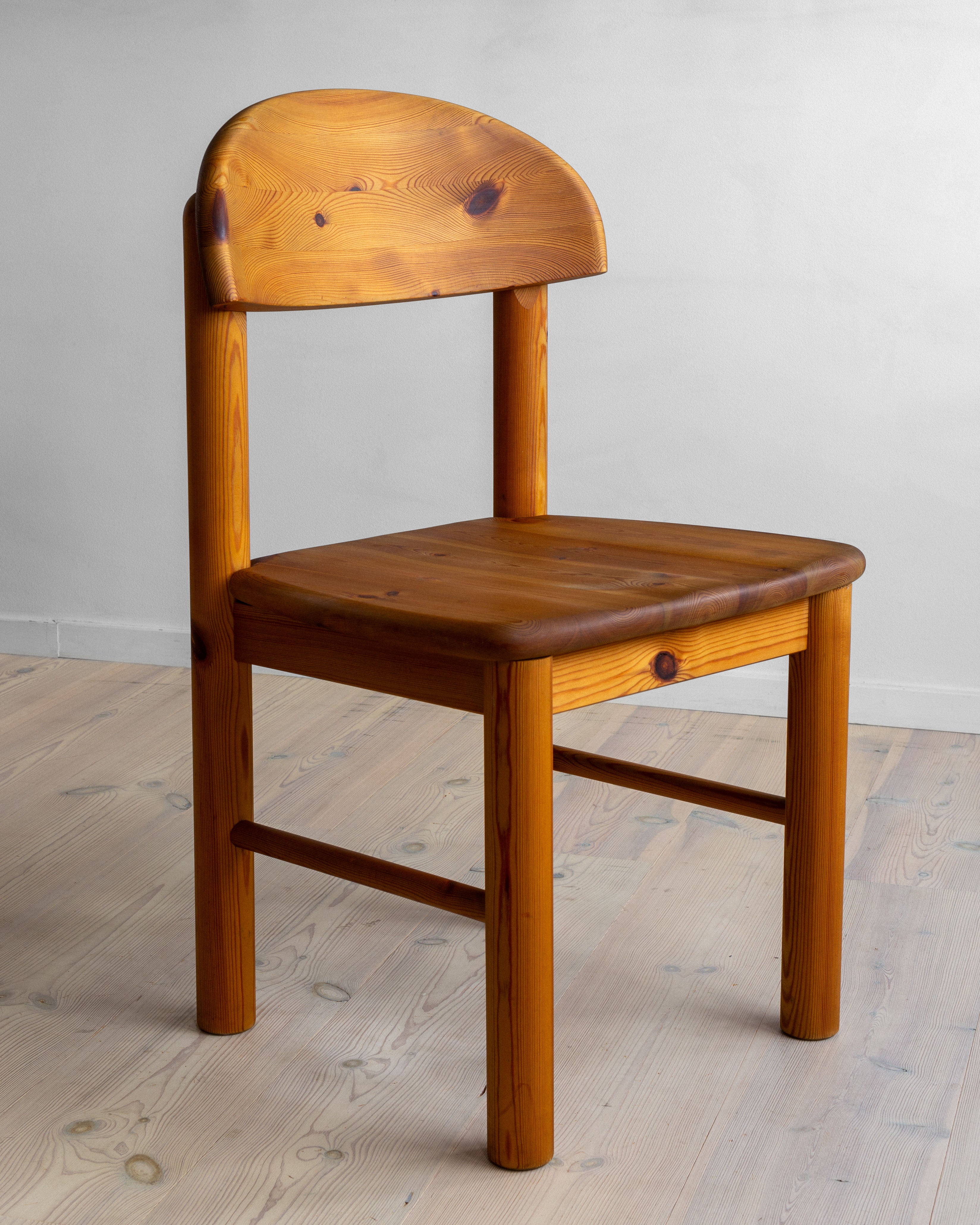 A set of 4 pine chairs by Rainer Daumiller, crafted in Denmark during the 1970s, is positioned on a light-colored wooden floor. The polished pine wood showcases the natural grain and warm, honey-toned finish that exemplifies solid pine furniture from Spigel. The background features a plain, light gray wall.