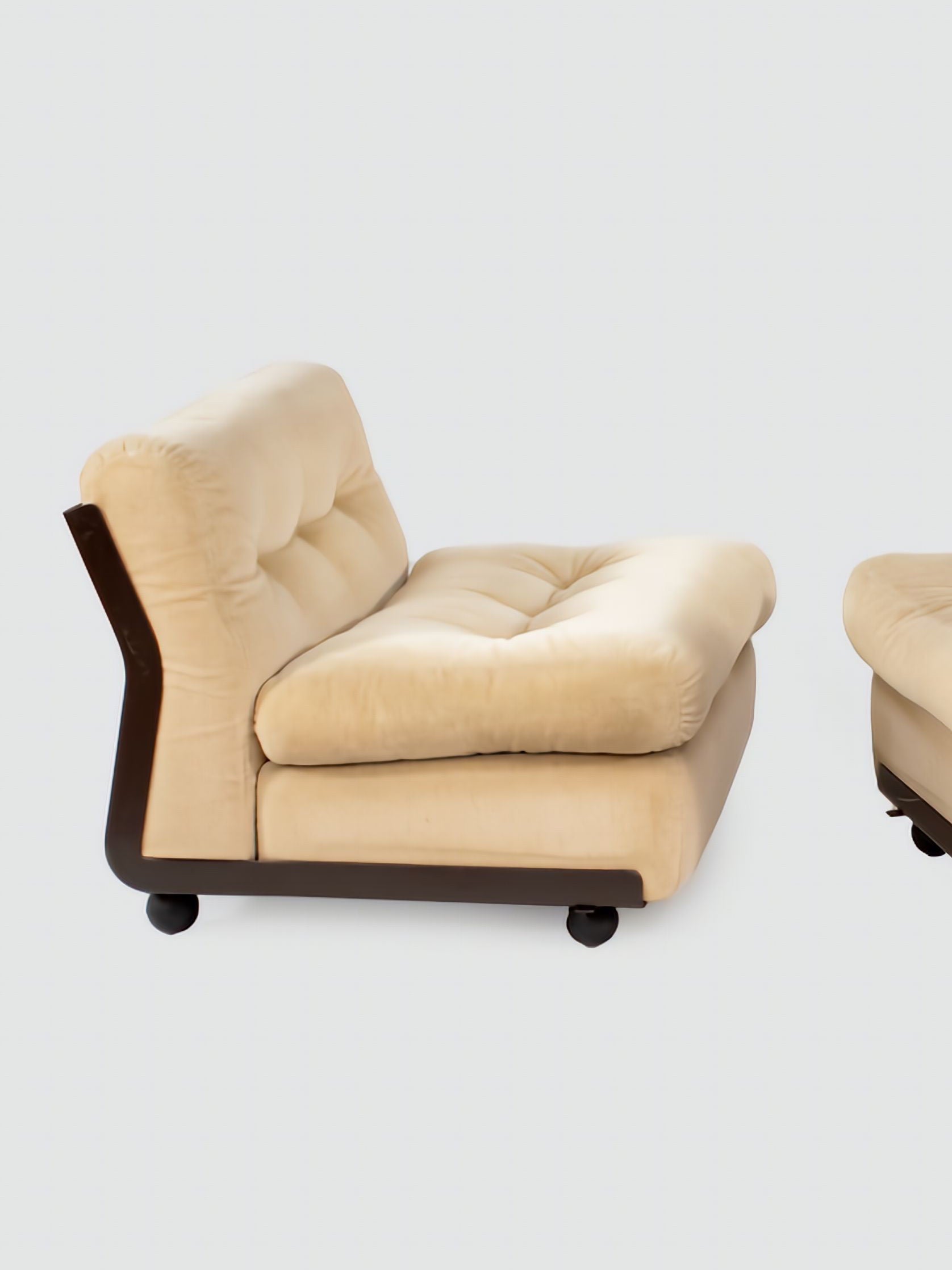 The pair of Amanta armchairs, designed by Mario Bellini in 1966 and available from RELIC LONDON, feature beige cushions with wooden frames and bases, and boast tufted backrests and seats. With their modern design, these chairs stand elegantly against a plain white background.