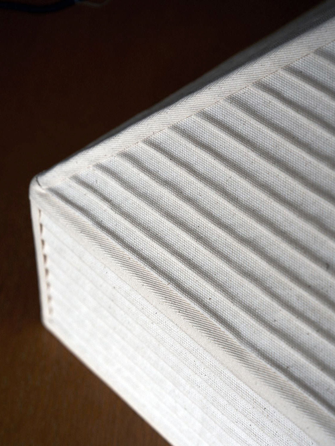 A corner of a white, canvas-covered Pyramid Hanging Lamp by Nassi Lamps with a textured, ribbed pattern is shown in close-up against a plain brown background. The rows on the lamp create a uniform, linear design.