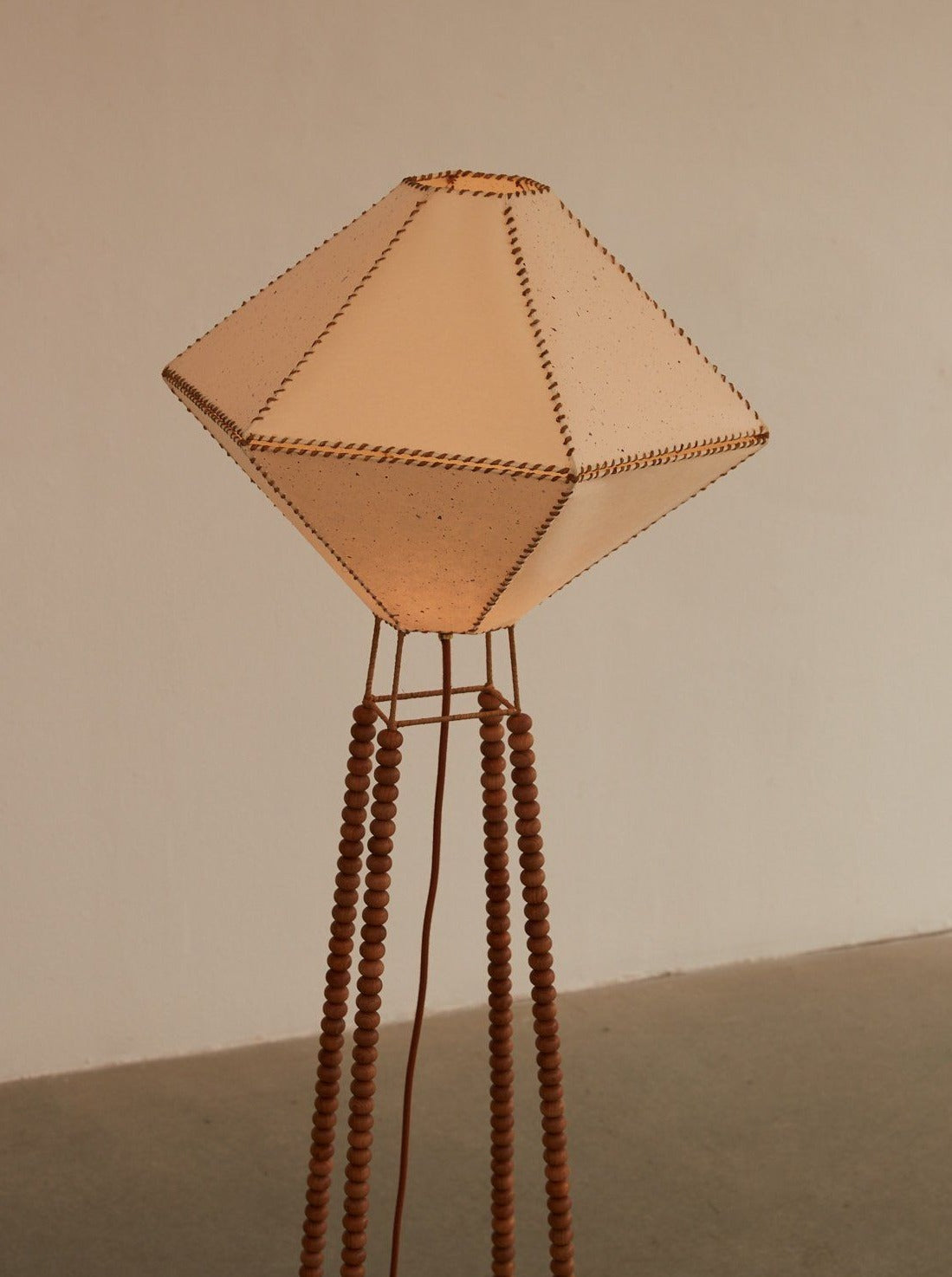 The USO Floor Lamp by Lana Launay is a piece of handmade lighting with a uniquely designed geometric shade, featuring beige fabric panels bordered with a woven texture. It stands on a slender, intricately beaded wooden base with multiple legs, combining artistic flair and contemporary style while using environmentally considered textiles.