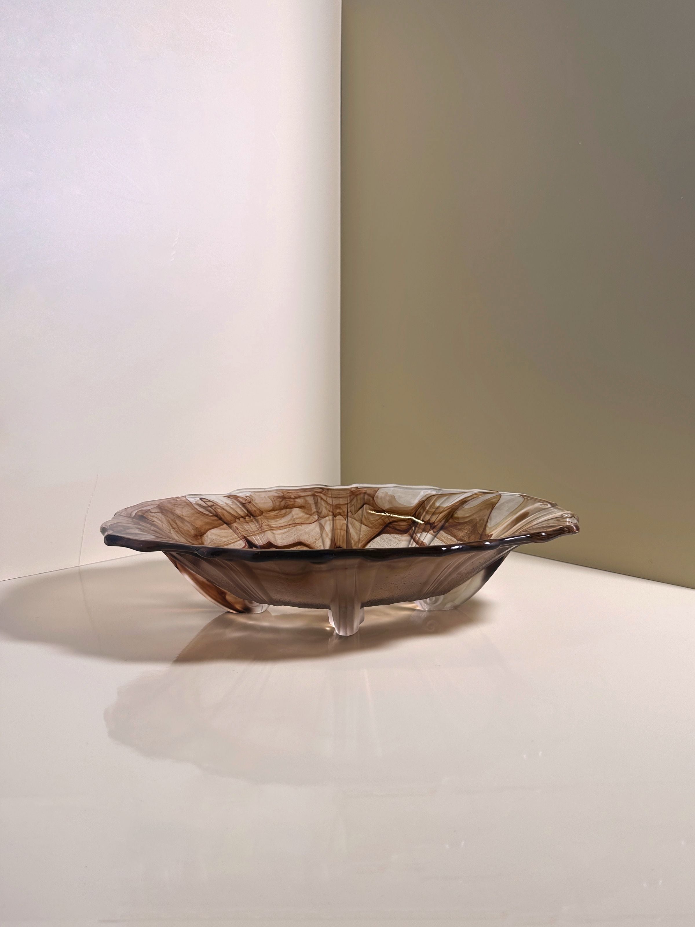 A delicate, translucent German Art Deco Cloud Glass Bowl circa 1930 by Dodo Vintage, featuring swirling brown patterns and a wavy edge, rests against a light-colored wall and surface. It casts a soft shadow with its elegant design, creating an artistic visual effect.