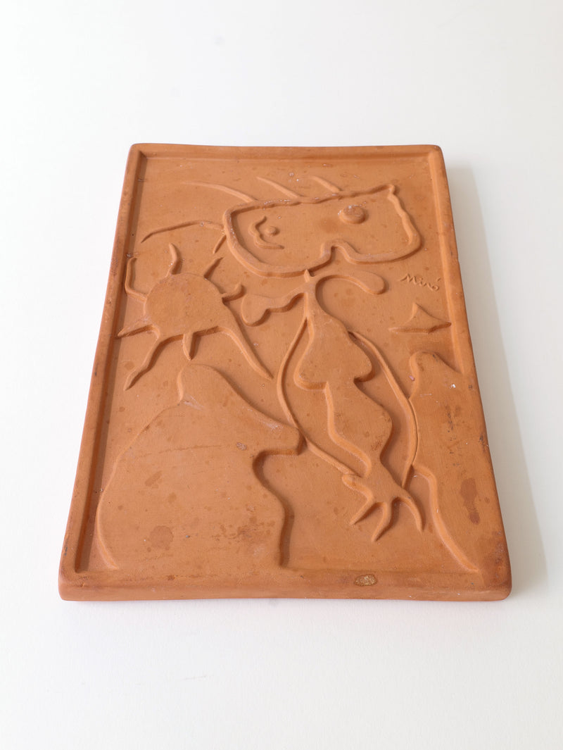 The Terracotta Relief Plaque Signed Joan Miró (1938) by Collection Apart features an abstract figure with elongated limbs and a bulbous head against a sunlit backdrop, set on an earth-toned rectangular slab, capturing the whimsical style of the Catalan surrealist artist.