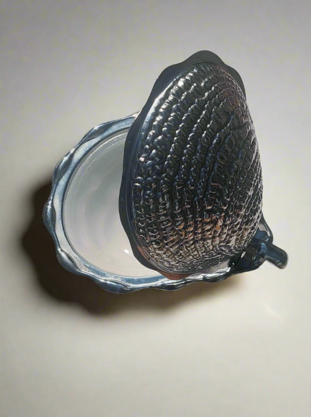 A Shell Butter Dish by Les Objoies, featuring a small ornate design with a textured, seashell-like lid, photographed on a marble surface. The lid is open, revealing an empty interior. The lighting casts a soft shadow beside the dish.