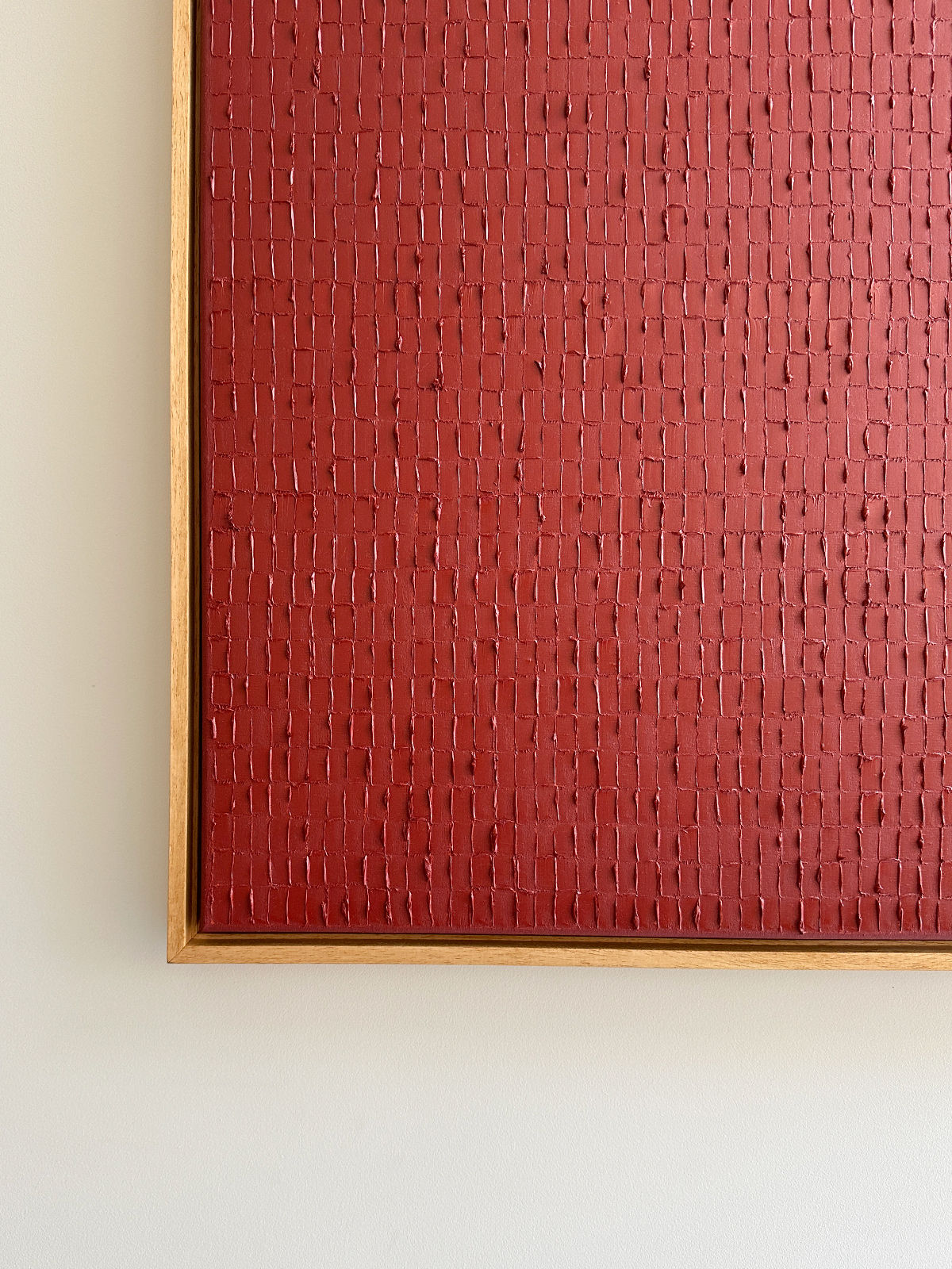 A rectangular artwork with a wooden frame features an intricate, textured pattern composed of small, uneven, raised red rectangles on a vibrant red background. Echoing the Dansaekhwa movement, the subtle woven appearance invokes mindfulness. The piece, "Indian Red - Oil on canvas" by Roi Elmaliah, is mounted on a white wall.