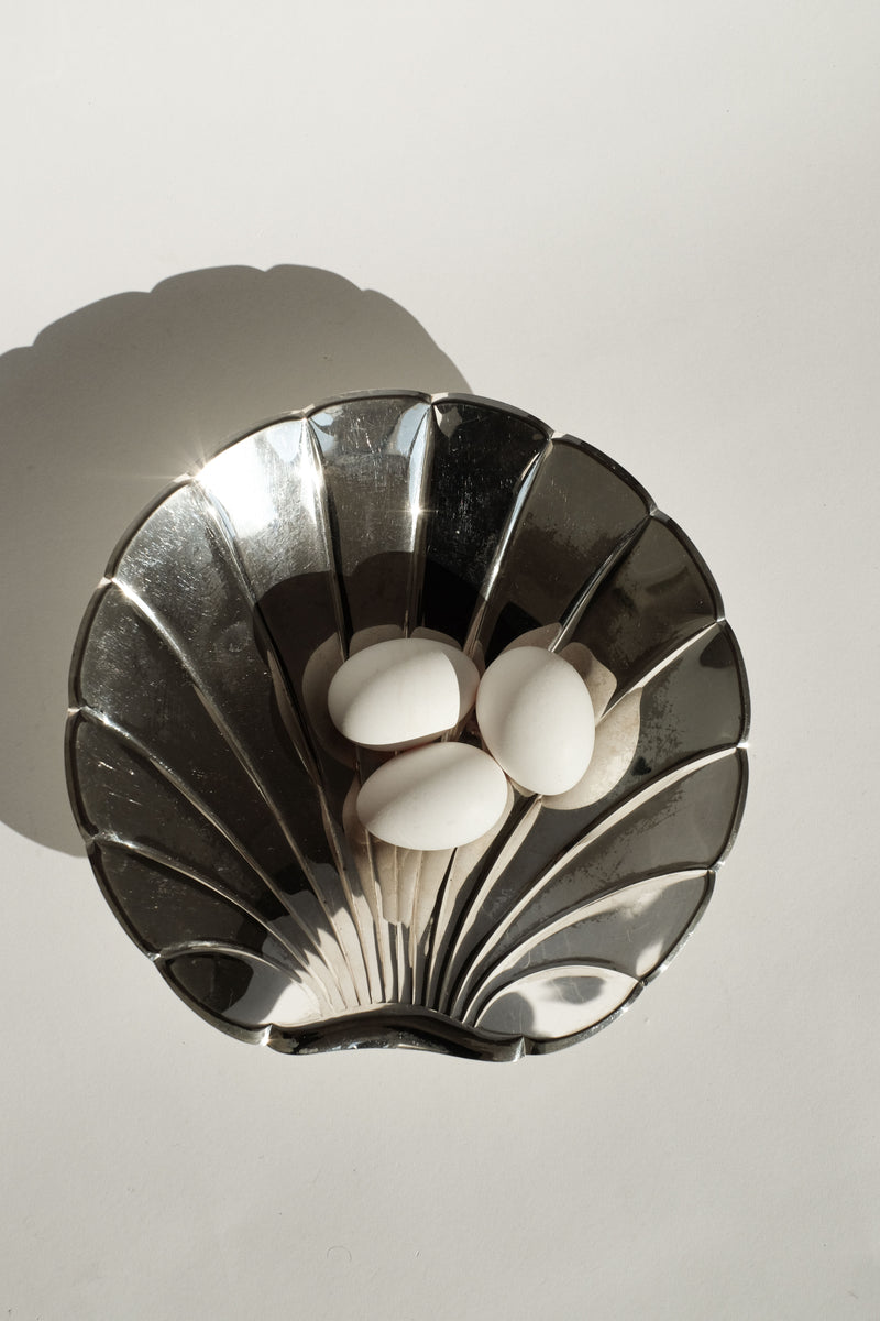 The Large Silverplated Shell Dish by septembre studios, with its natural patina glow, holds four smooth white stones. Its vintage design casts a soft shadow on a light surface as sunlight dances across its shiny contours.
