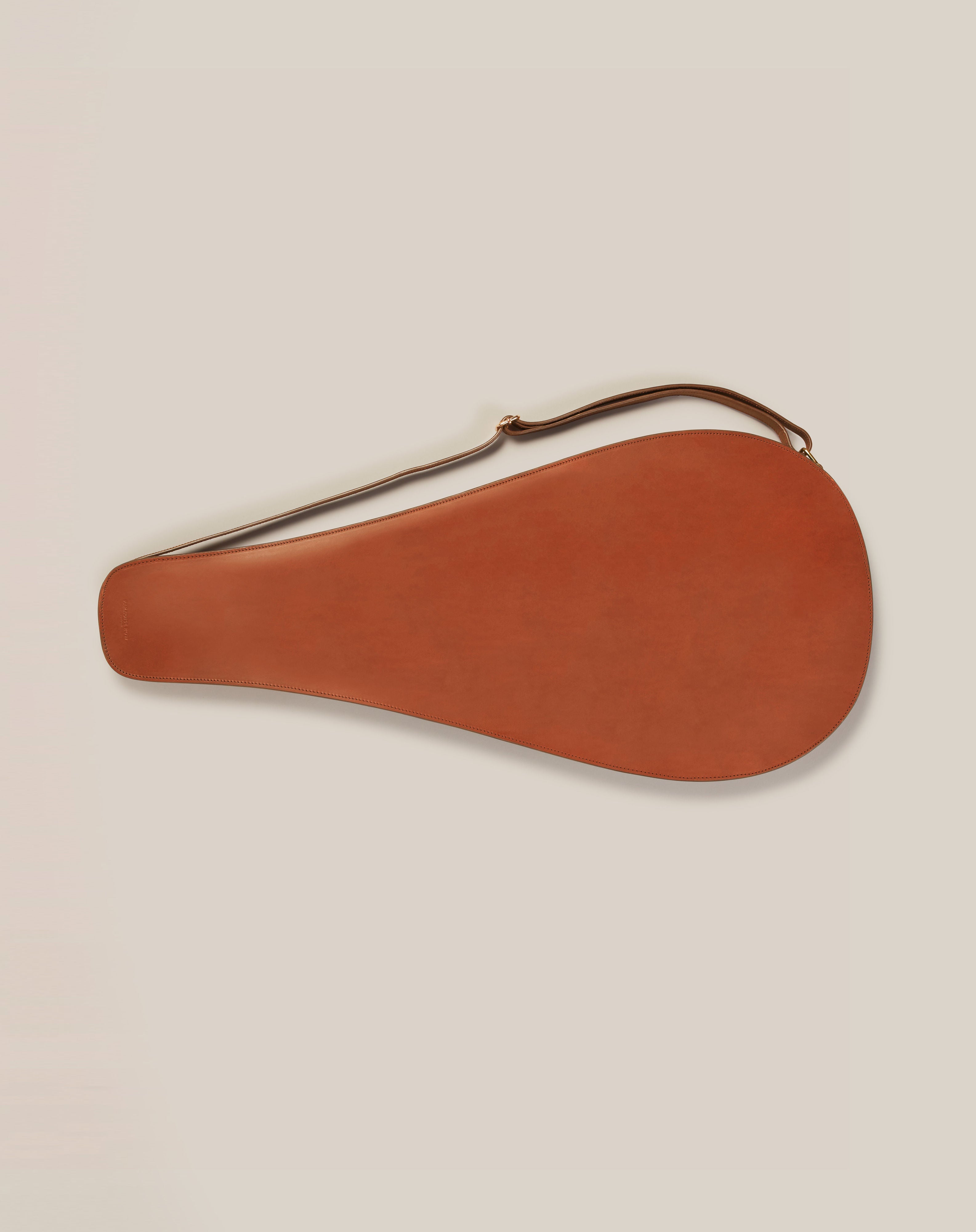 The Leather Tennis Racket Cover by Paradise Row, a teardrop-shaped brown design with a long strap, exhibits bespoke British craftsmanship. Made from sustainable vegetable-tanned leather, it elegantly rests against a light background.