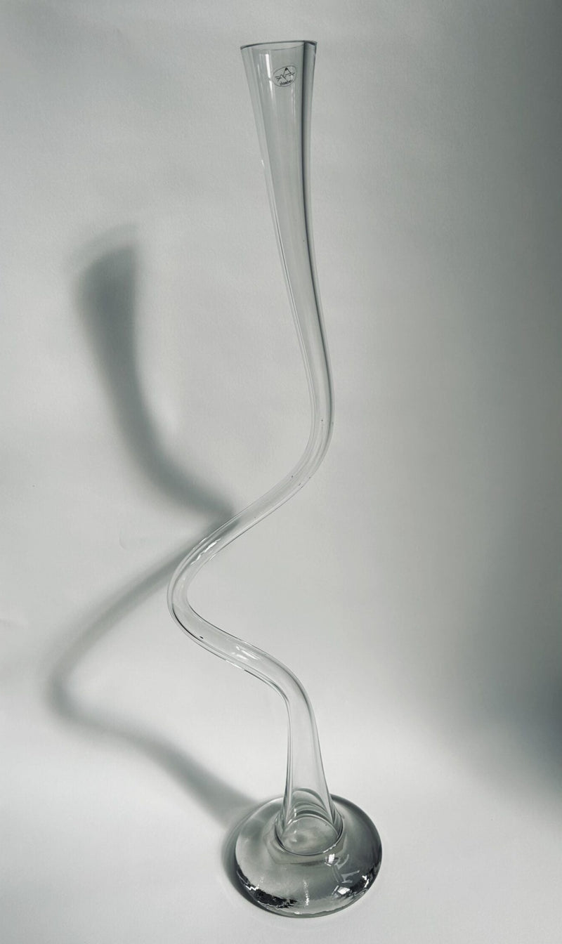 The Swirl Vase (large) by Flock Studio is a tall, clear glass vase featuring a twisted, elegant design that makes it a striking decorative object. Handmade in the Netherlands, it stands on a round base and casts a subtle shadow on a light background.