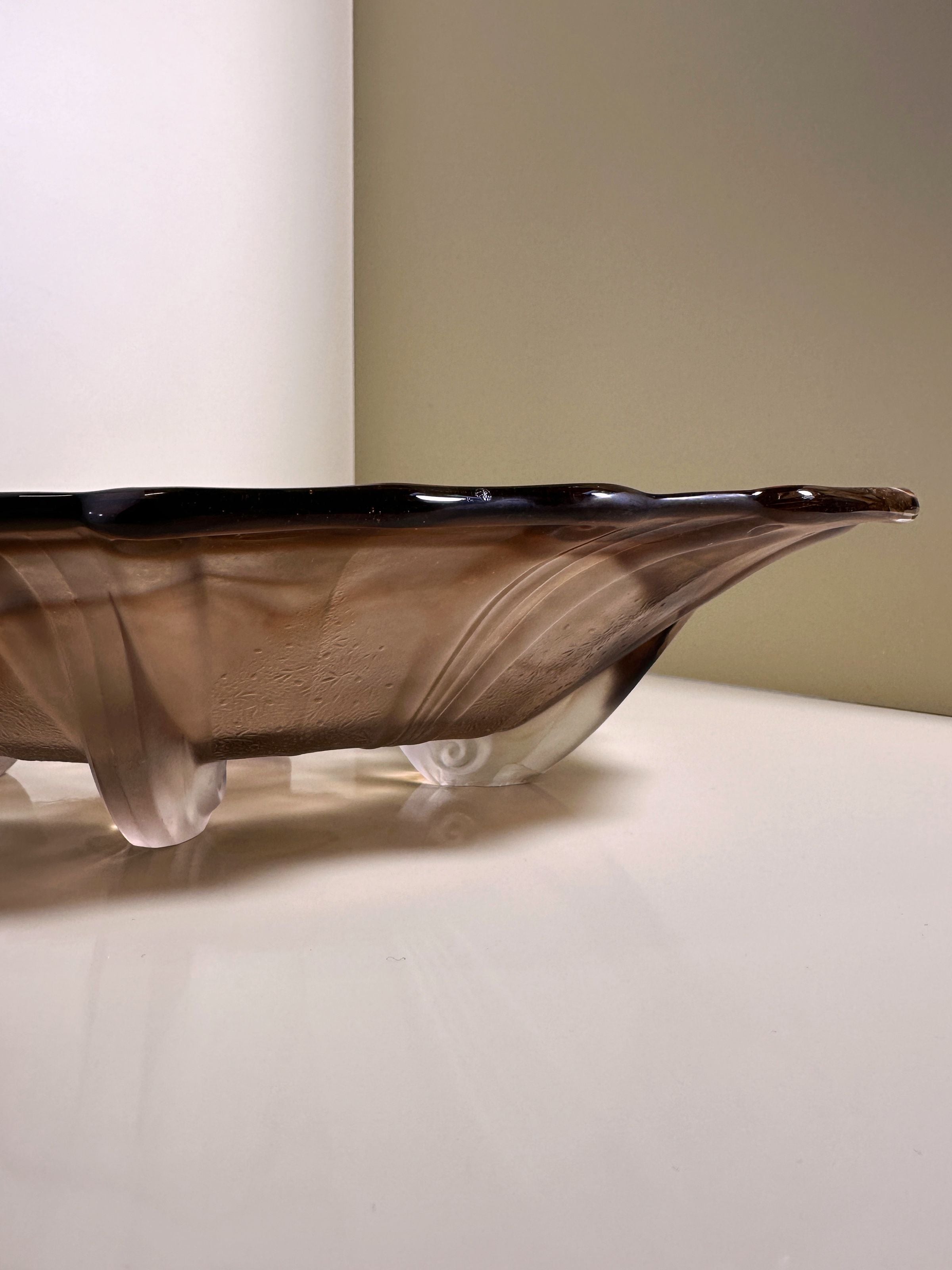 A sleek Art Deco glass bowl from Dodo Vintage, circa 1930, features an elongated design with a dark smoky hue and fluid, wave-like contours. Its surface casts elegant shadows and reflections on white.