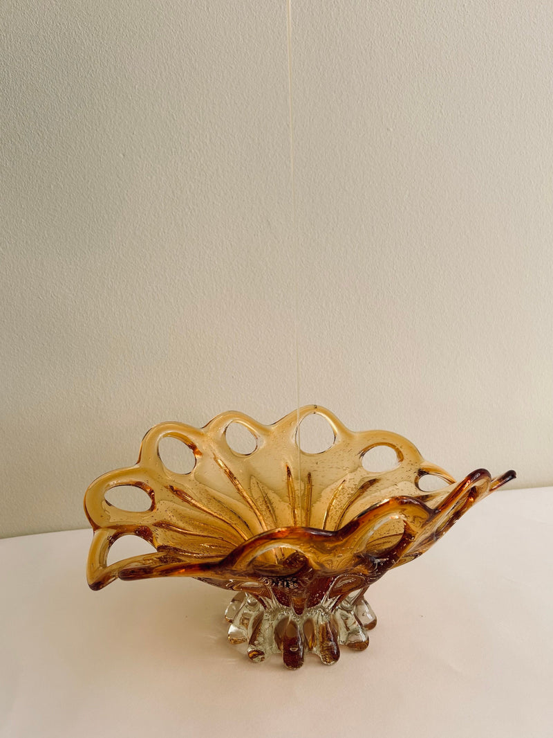 A Murano Glass Amber Bowl by Flock Studio, featuring a wavy, scalloped edge and intricate cut-out designs, handcrafted with Italian glass artistry. It is displayed on a white surface against a light-colored background.