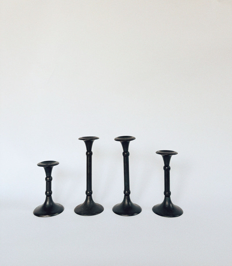 The Huerto Set of 4 Cast Iron Candleholders features black candlesticks with rounded bases and slim stems, arranged against a light background. Their simple design captures the timeless elegance of vintage candlesticks.