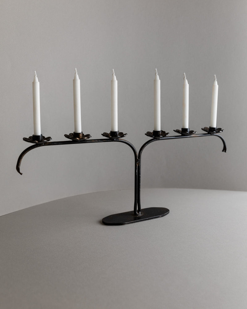 The Iron Candleholder from Bottega Jacobs, Italy 1960s, is a black metal piece with an L-shaped design that elegantly holds five unlit white taper candles. Its minimalist brutalist style complements any neutral gray surface against a gray background.
