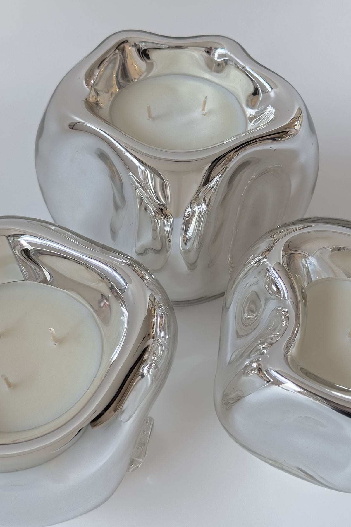 Three HOMA Lumi Candles, crafted with silver mirror glass and each featuring a white two-wick candle, are arranged closely on a light surface. Their smooth, rounded design elegantly reflects light and surroundings.