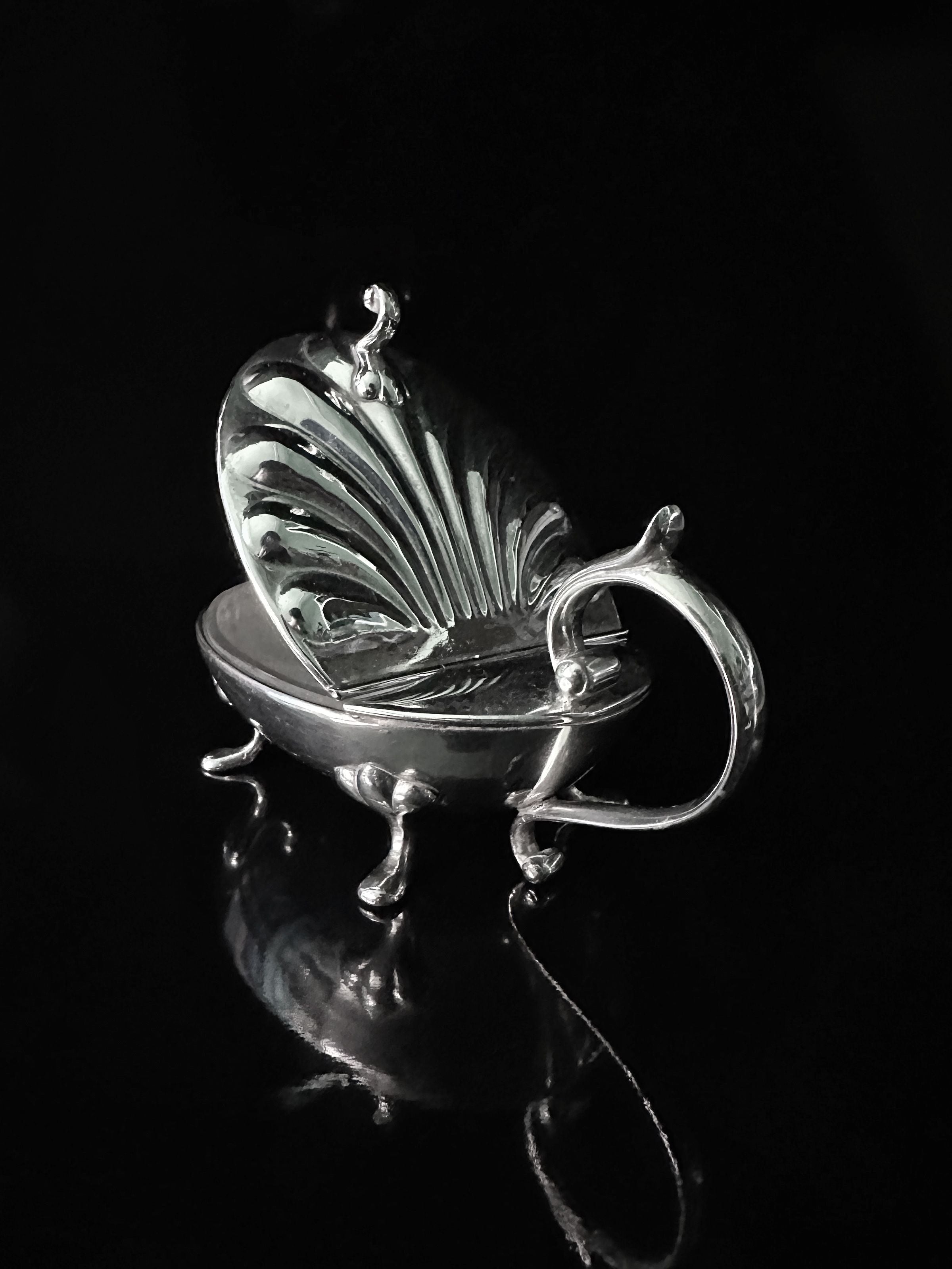 The Dodo Vintage Silver-plated Caviar Frog 1950s, with ornate detailing and a smooth interior perfect for caviar, resembles a clam shell. It rests elegantly on four small legs atop a reflective black surface, casting a subtle shadow.