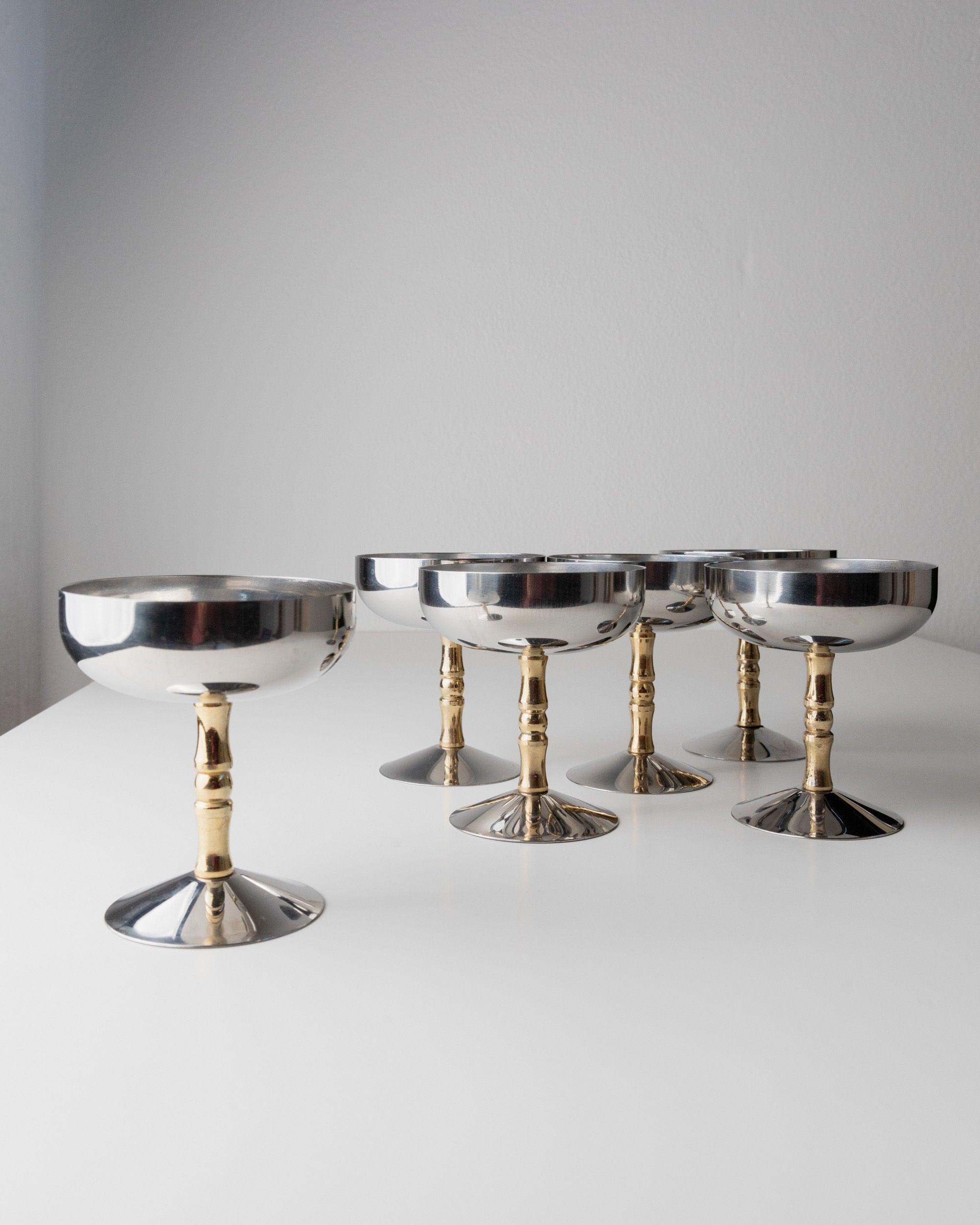 Six Bottega Jacobs 6 Stainless Steel Champagne Coupes 70s with gold-plated handles are artfully displayed on a white surface against a simple background, showcasing their minimalist and elegant style.