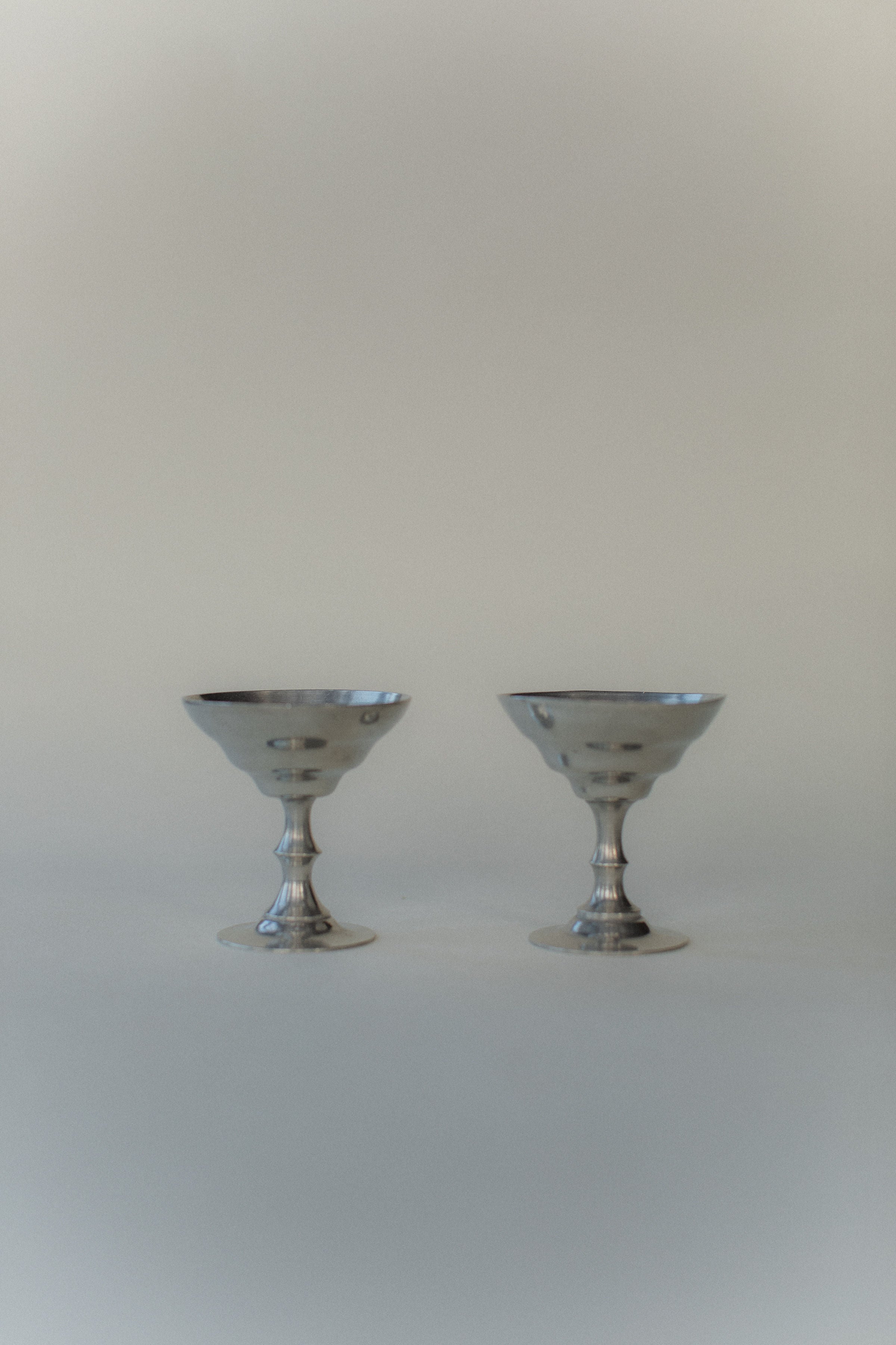 Two silver vintage-style ice cream cups from Out For Lunch, with scalloped edges, stand side by side on a smooth light gray surface. Reminiscent of Art Deco style, the softly blurred background enhances the minimalist elegance of the scene.