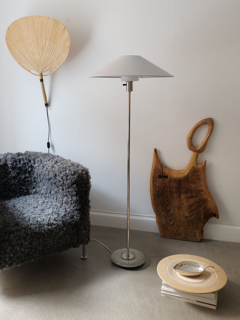 A cozy living room corner with a textured gray armchair, a Collection apart Deco Floor Lamp Harald Notini 1940s with a gray shade, a wall-mounted fan, a large wooden cutting board, and a small table holding books and a bowl.